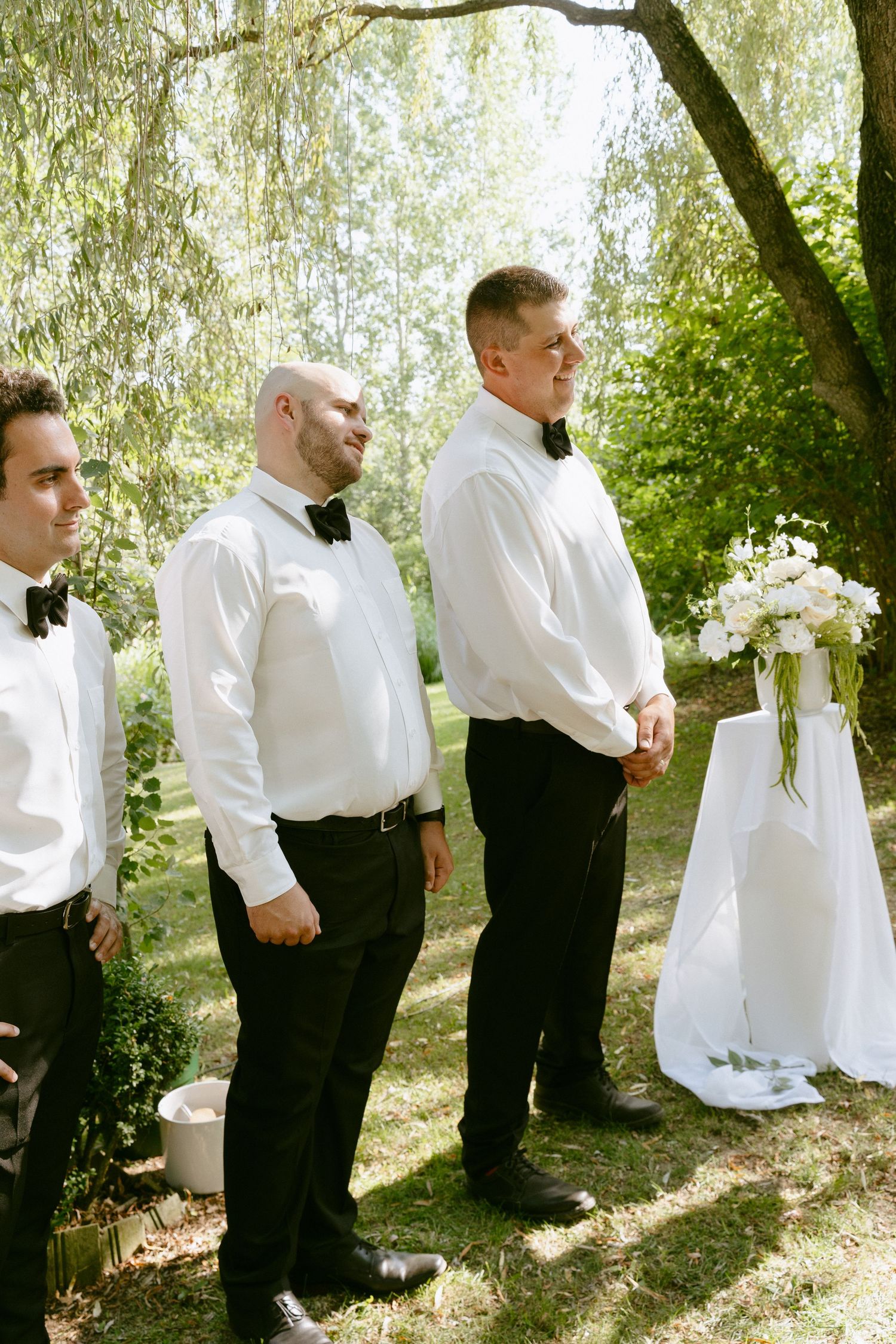Three groomsmen in white shirts and bow ties stand at outdoor wedding altar.