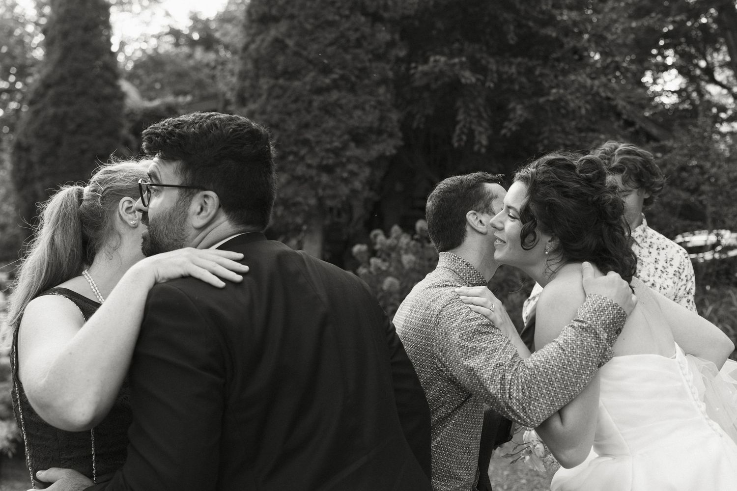 Two couples share a romantic kiss outdoors at a wedding celebration in black and white photography.