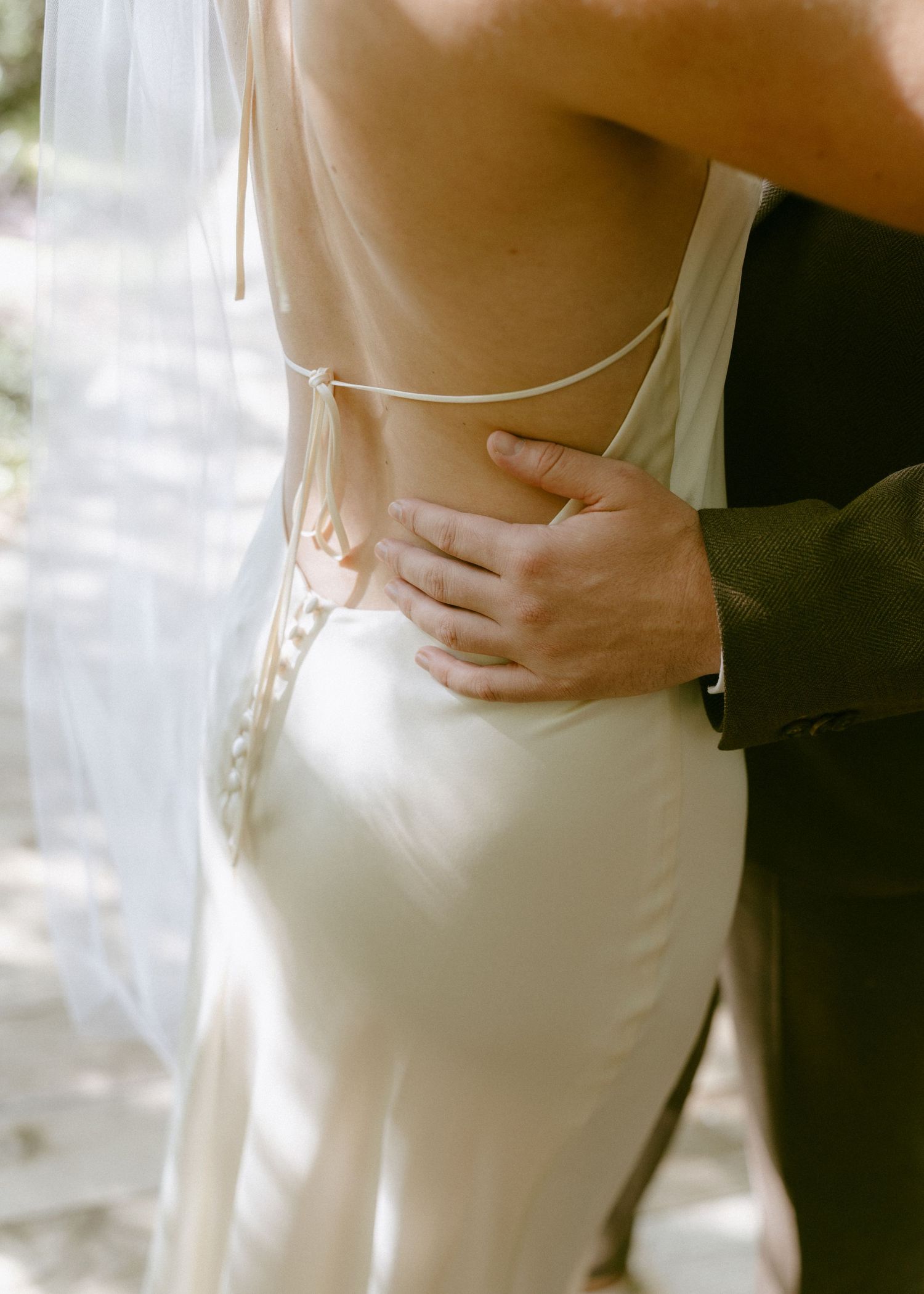 Close-up of groom placing hand on bride's back, highlighting the elegant open-back detail of her satin wedding gown.