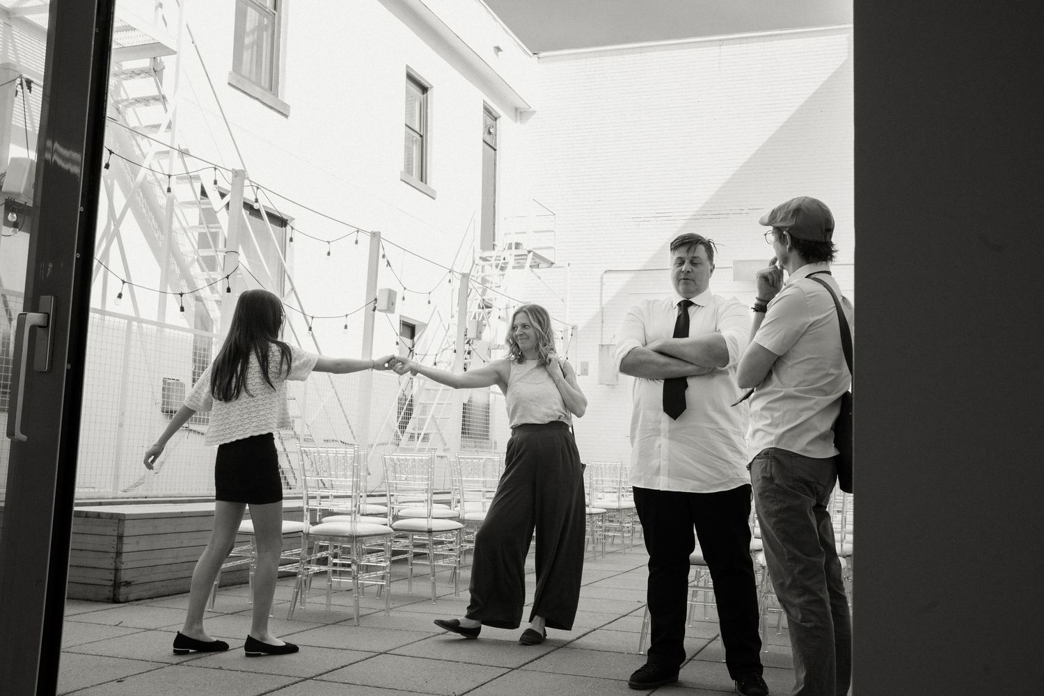 Black and white photo of four people socializing in an outdoor courtyard with string lights and chairs.
