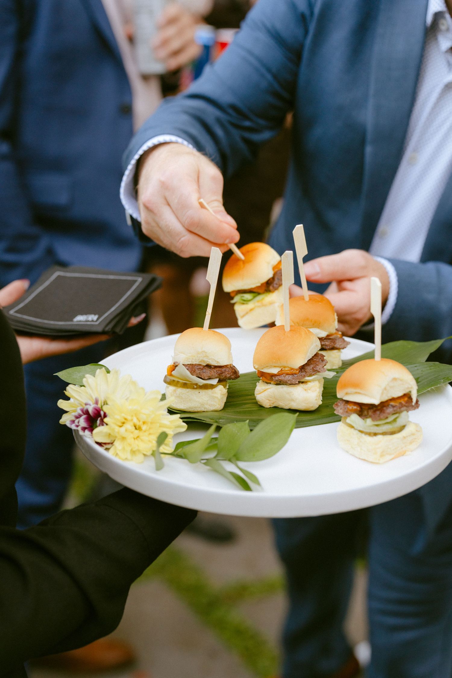 Guest reaches for a mini slider from a white tray of gourmet burgers garnished with flowers at a wedding.