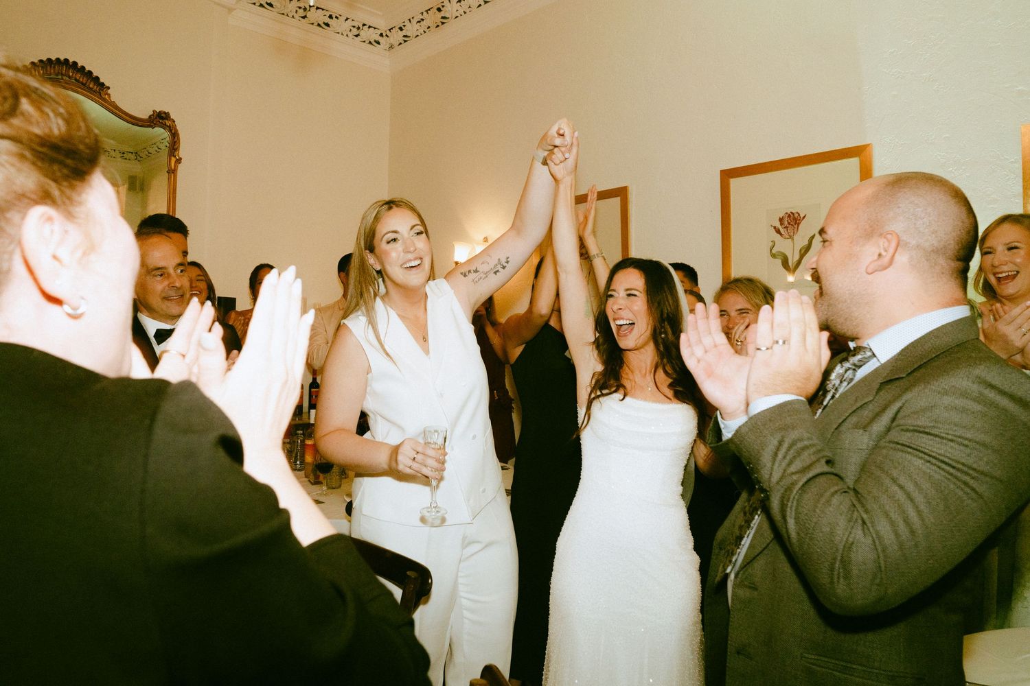 Two brides in white raising hands together while wedding guests applaud in celebration.