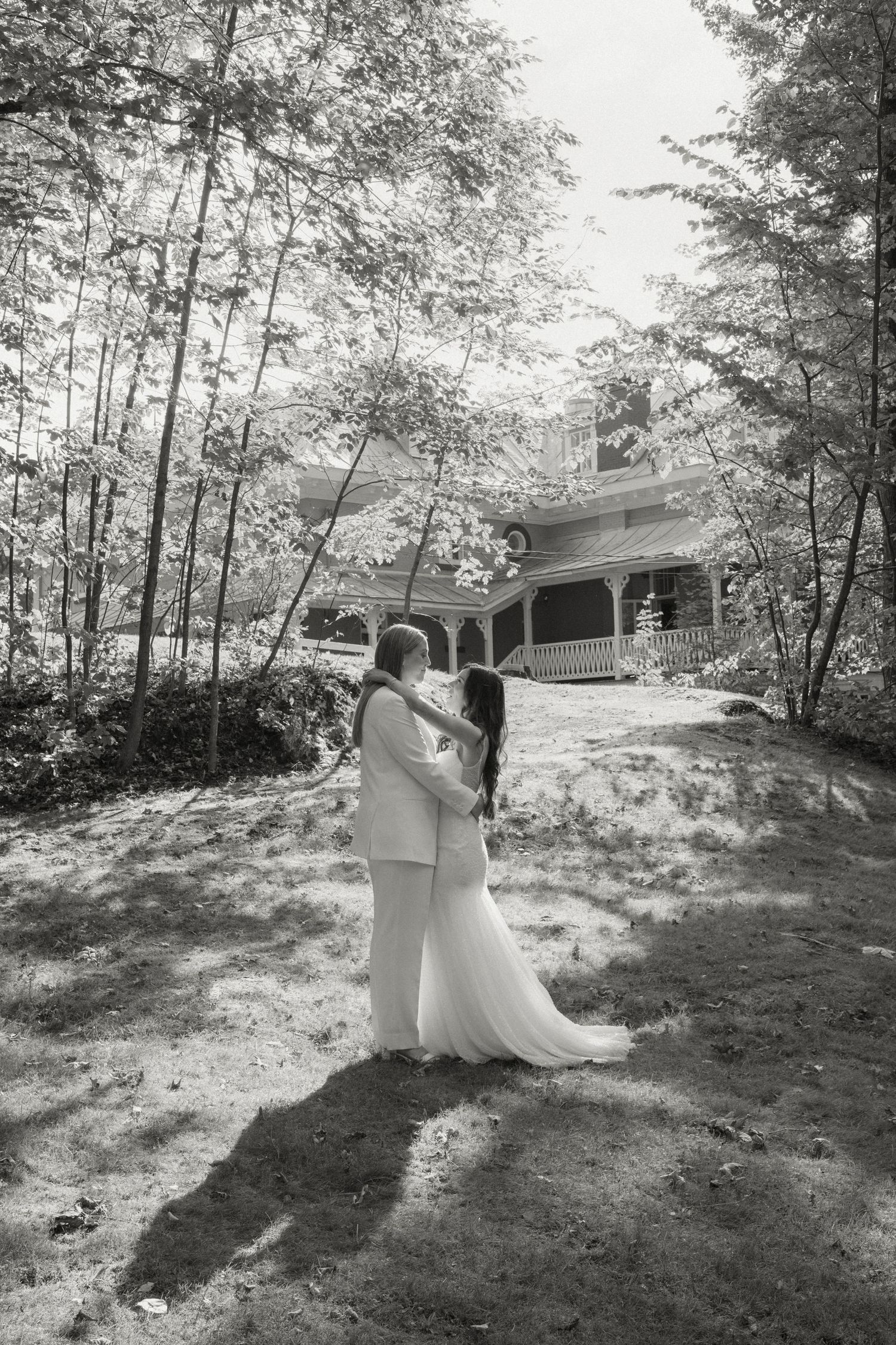 Two brides embrace outdoors in black and white photo with historic venue behind them.