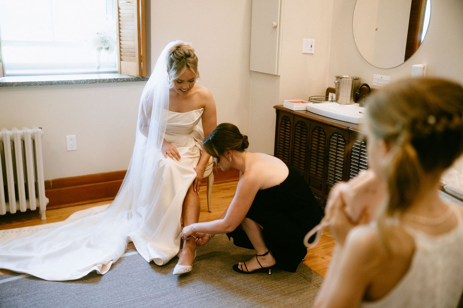 Bridesmaid helping bride with shoes while another bridesmaid adjusts her dress in a bright room.