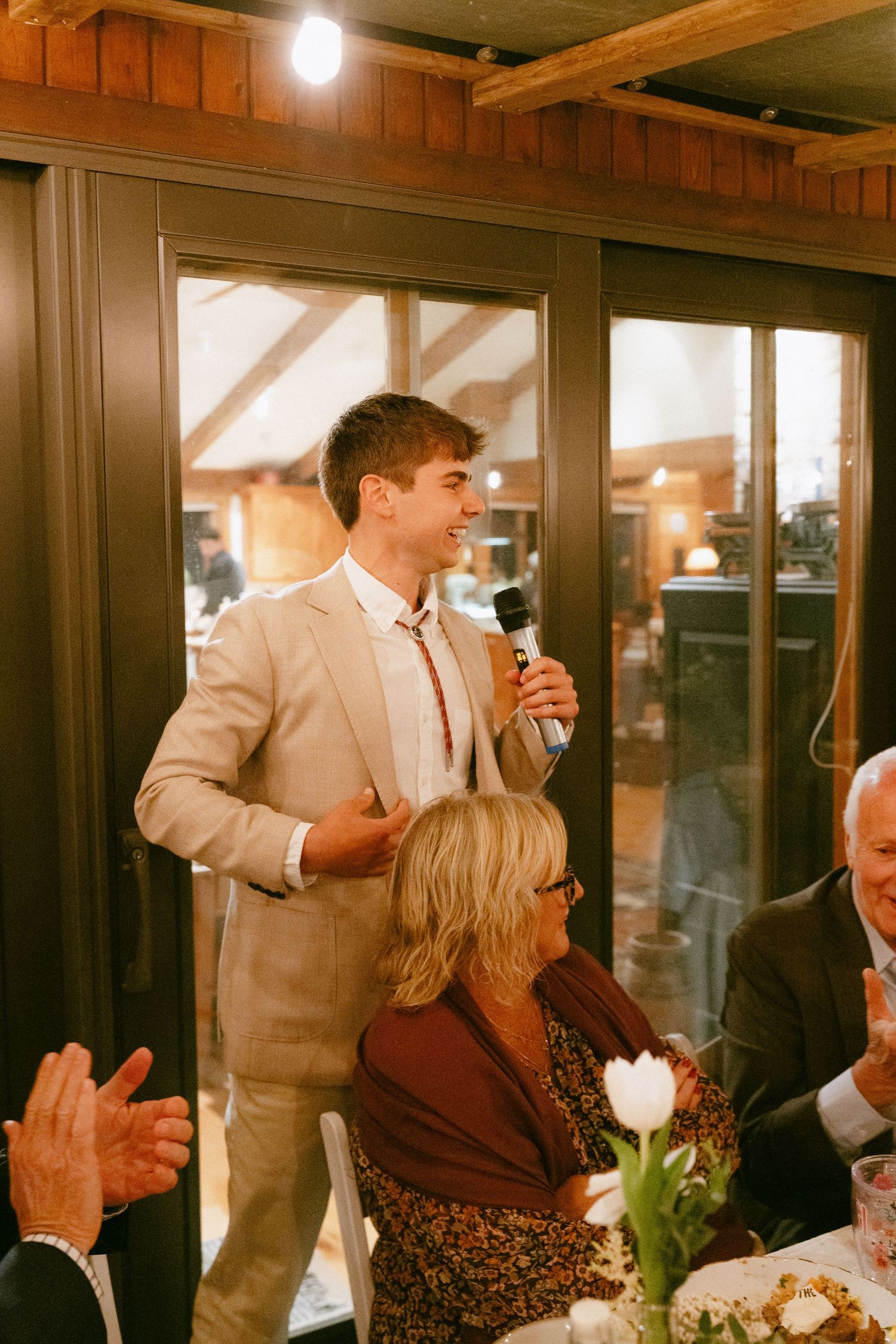 Young man in tan suit giving a speech with microphone at elegant wedding reception dinner.