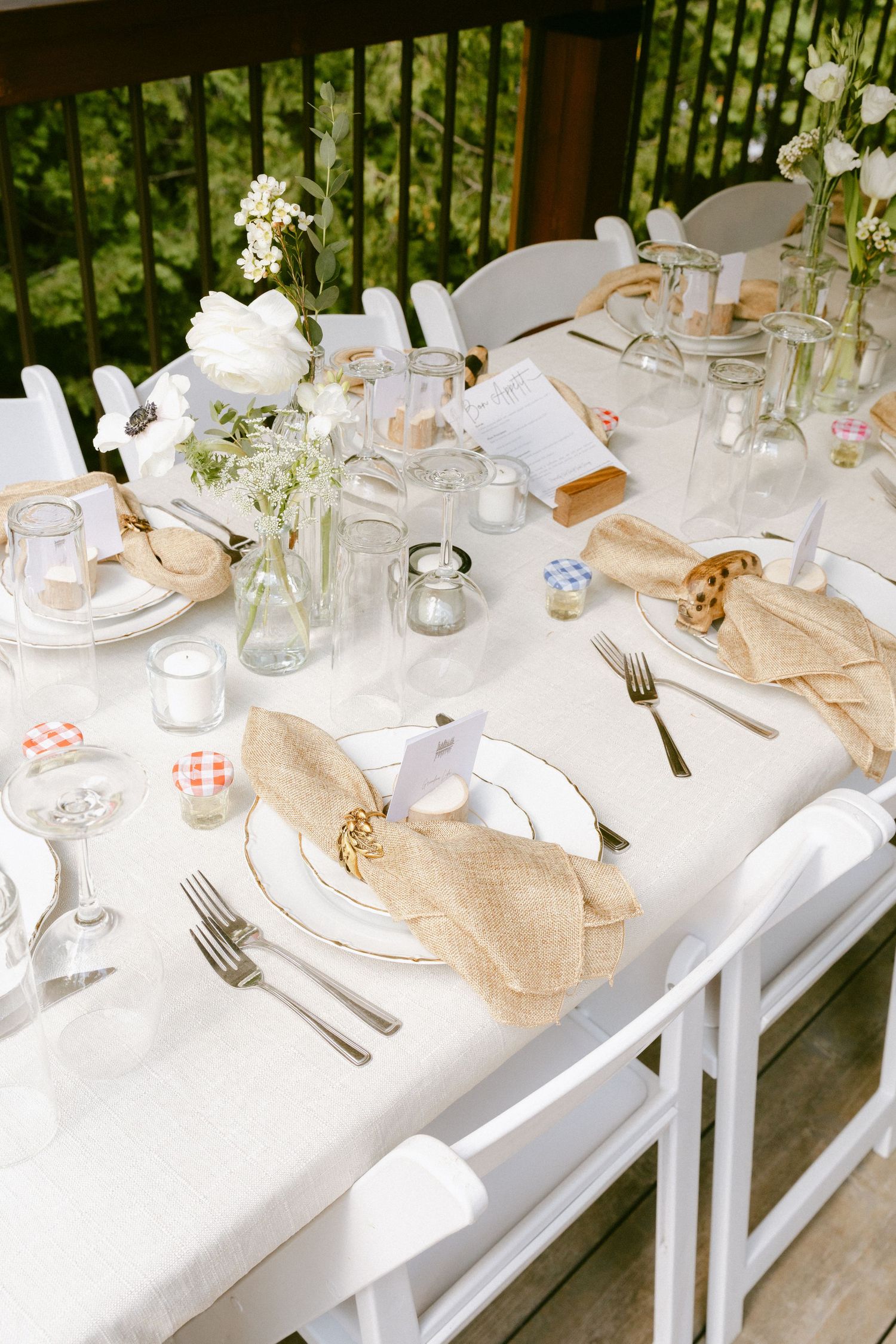Elegant outdoor wedding reception table set with burlap napkins, white florals, glass bottles, and candles on white linen.
