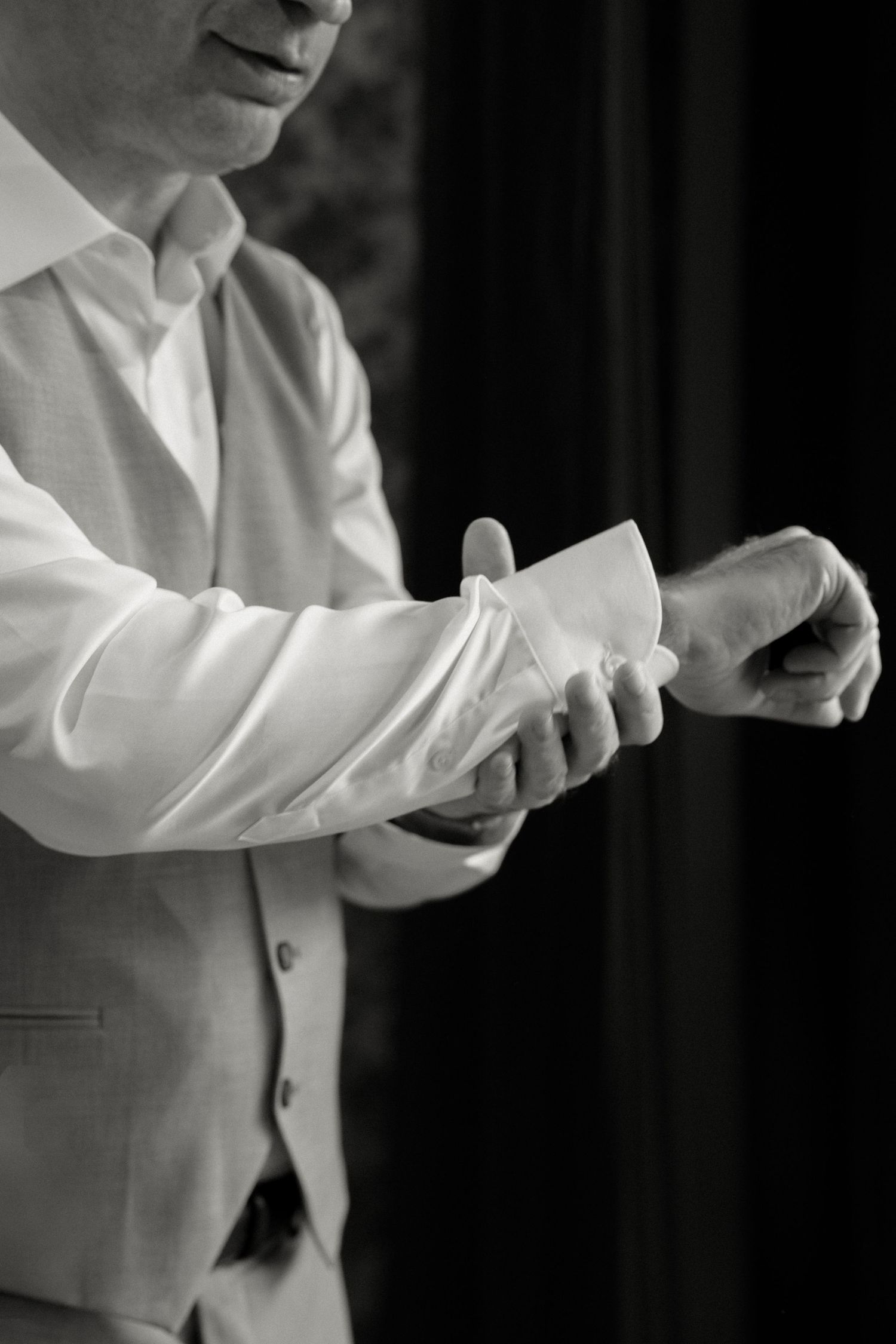 Black and white photo of groom in gray vest fastening cufflinks on white dress shirt.