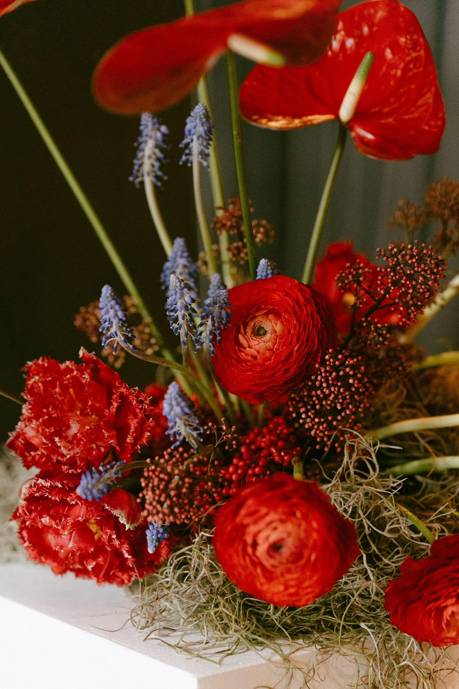 Vibrant red floral arrangement featuring anthuriums, ranunculus, tulips, and blue muscari with Spanish moss accents.