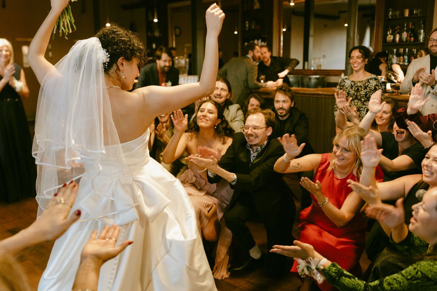 Excited wedding guests reach toward the bride during a lively indoor reception celebration.