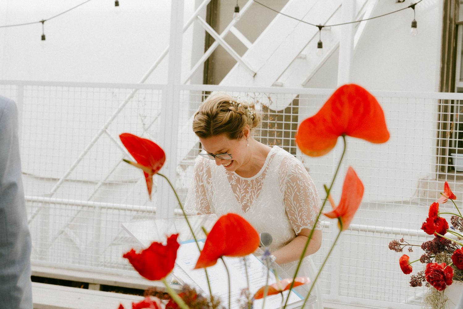Smiling bride in lace wedding dress surrounded by vibrant red poppy flowers at outdoor wedding venue.