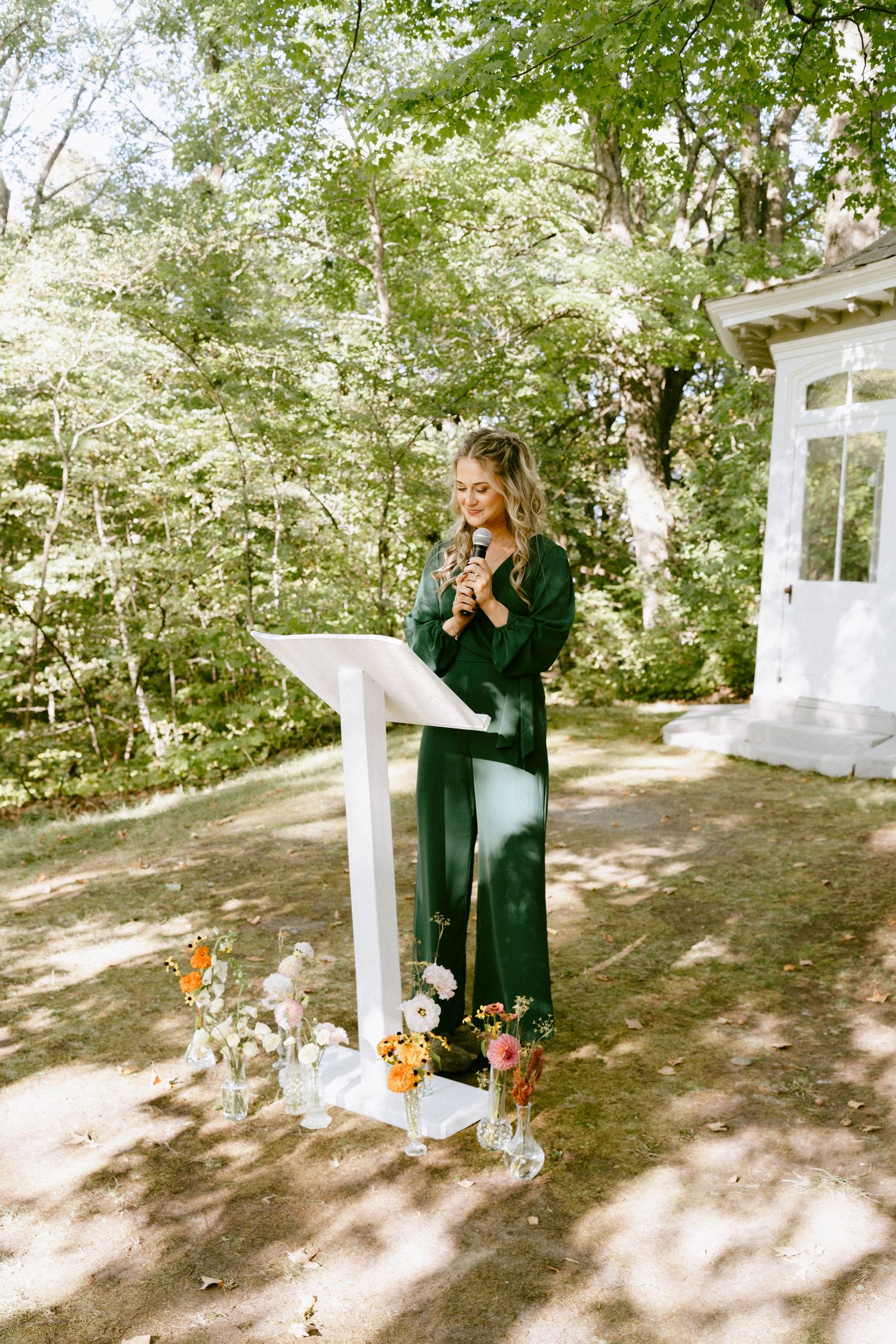 Female officiant in green dress speaks at white podium during outdoor garden wedding ceremony.