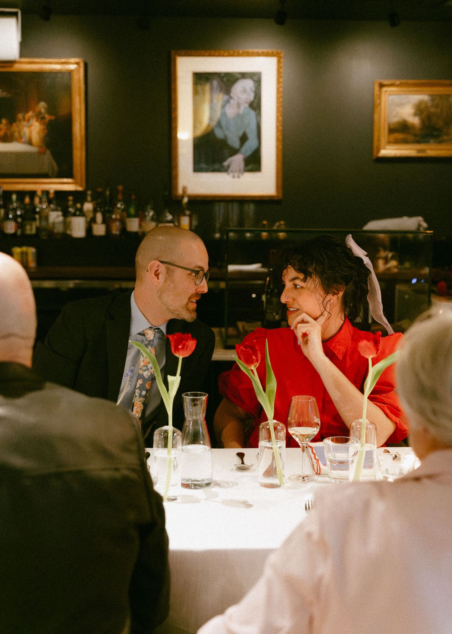 Bride in red dress and groom in suit share an intimate moment at their wedding reception dinner table.