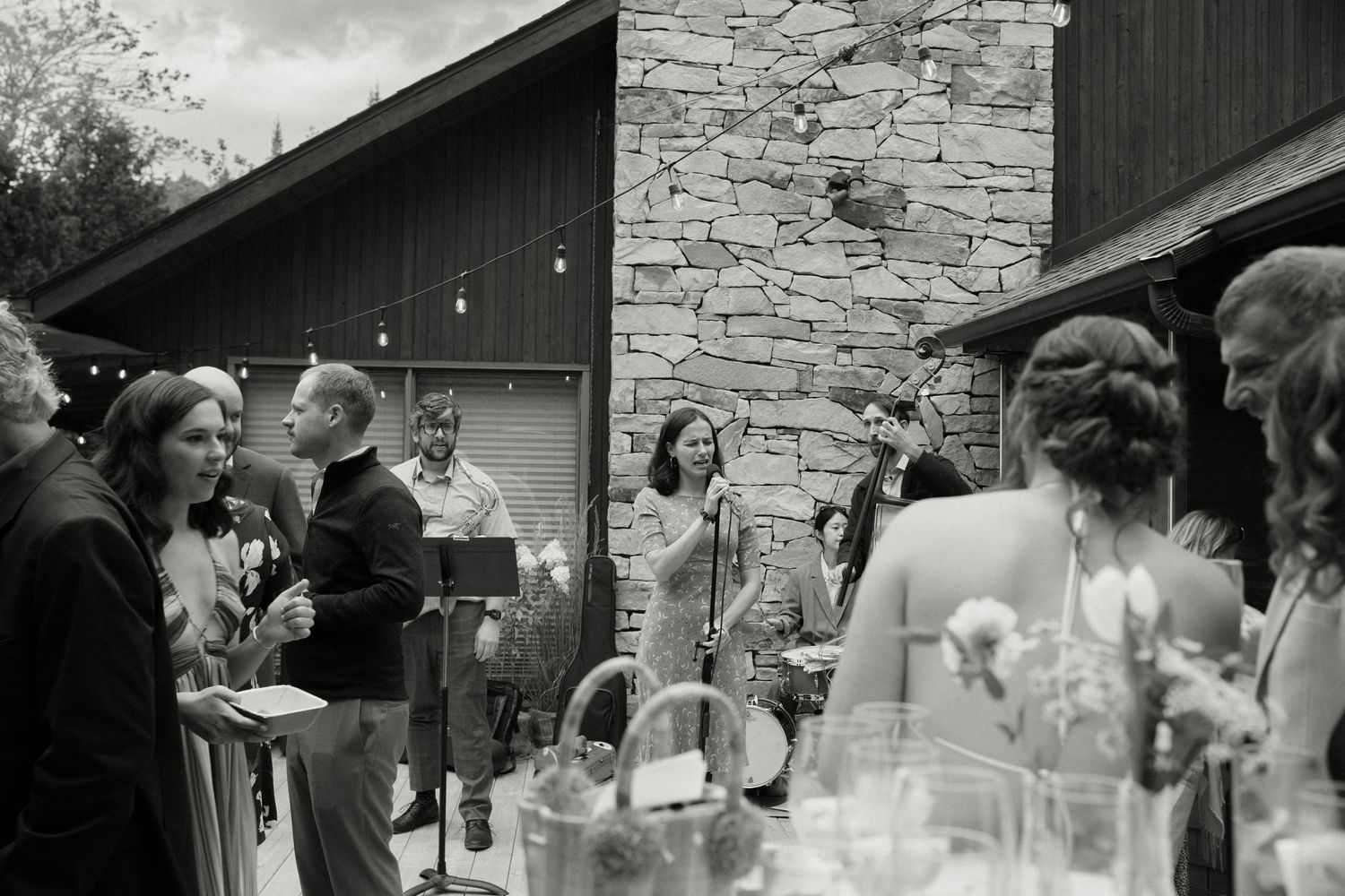 Black and white photo of wedding guests mingling outdoors near stone building with string lights.
