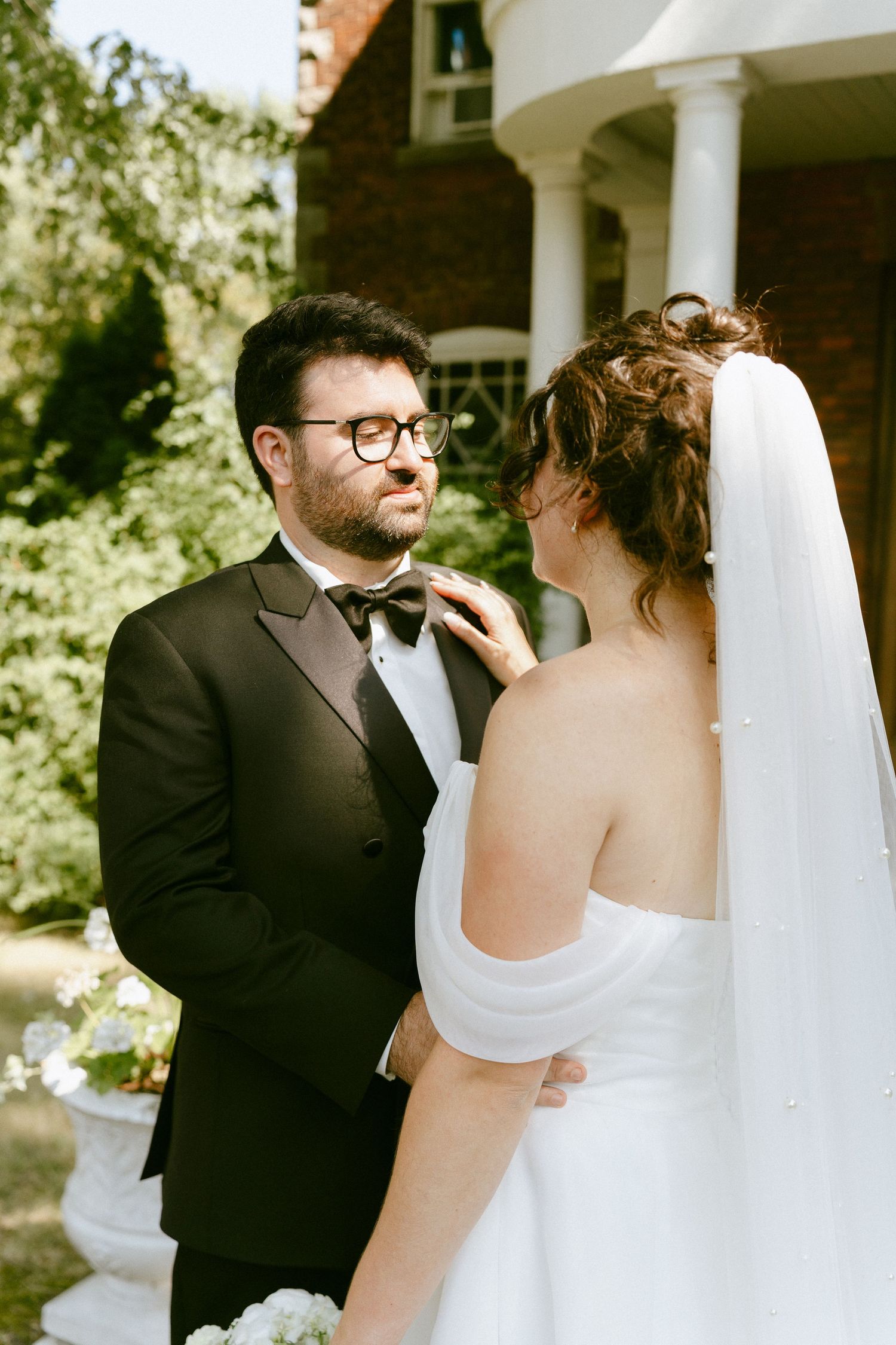Groom in black tuxedo gazes at bride as she adjusts his bow tie outdoors.