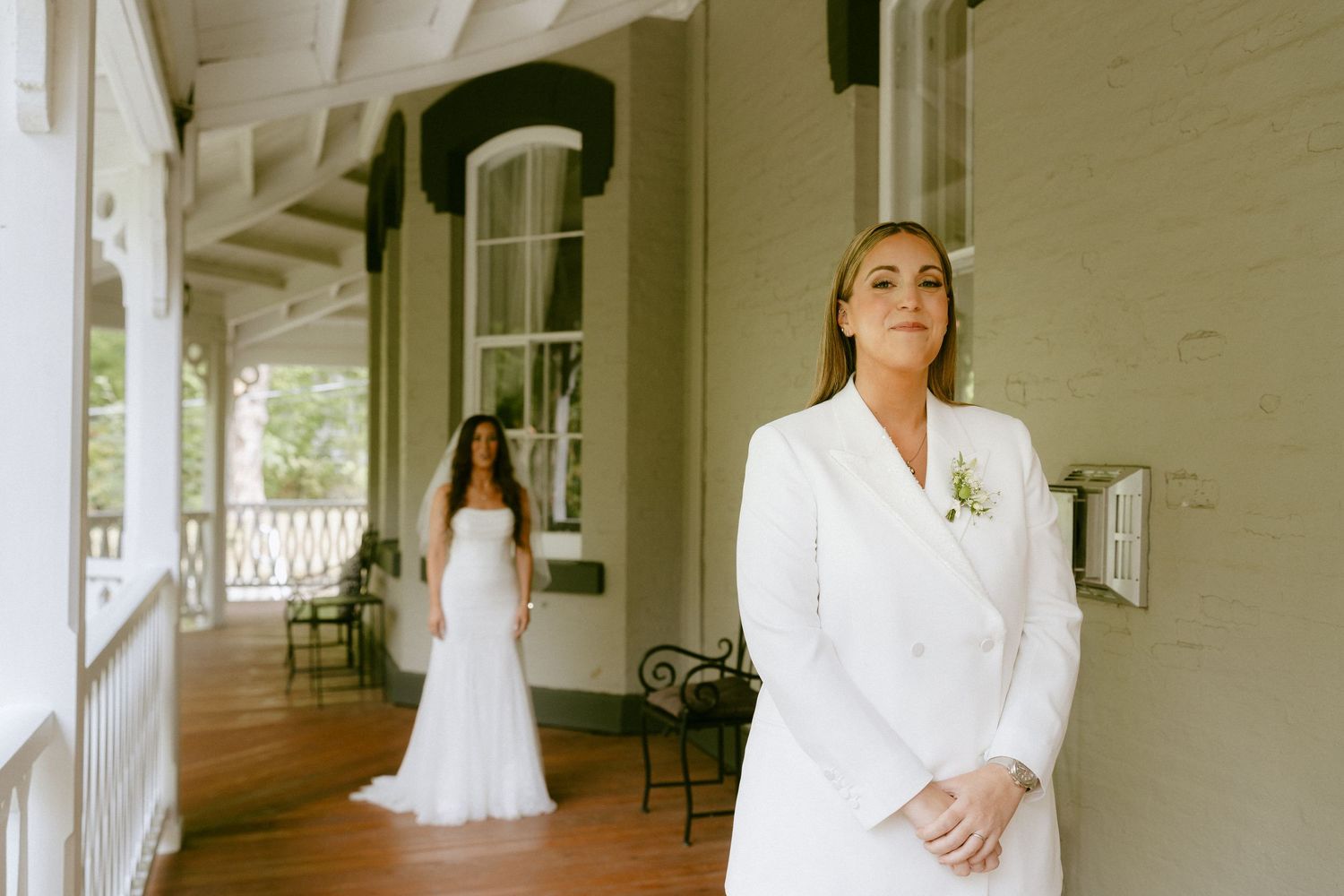 Two brides in white preparing for first look on elegant porch.