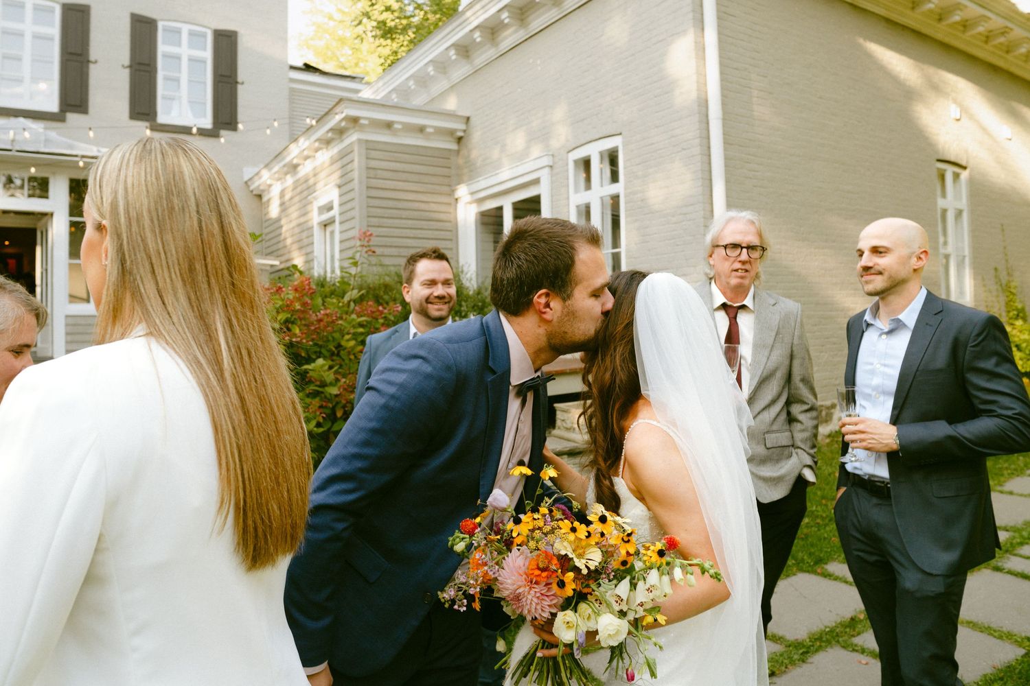 Wedding couple sharing a kiss while guests celebrate outdoors at a garden wedding reception.
