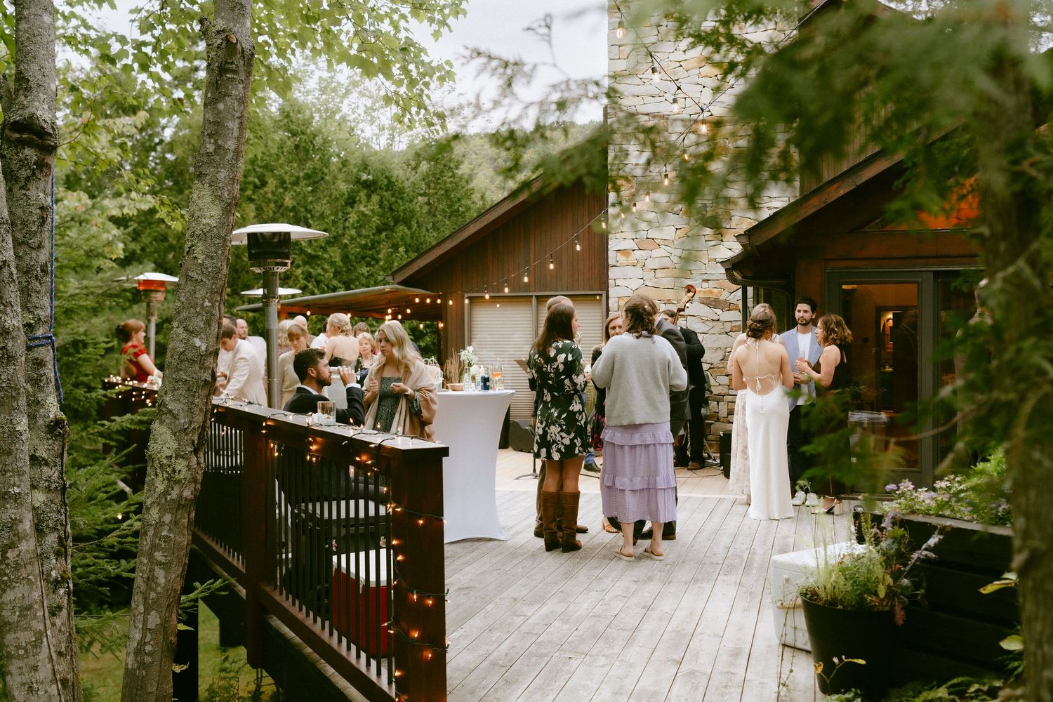 Guests mingling on wooden deck at intimate outdoor wedding reception surrounded by trees and string lights.