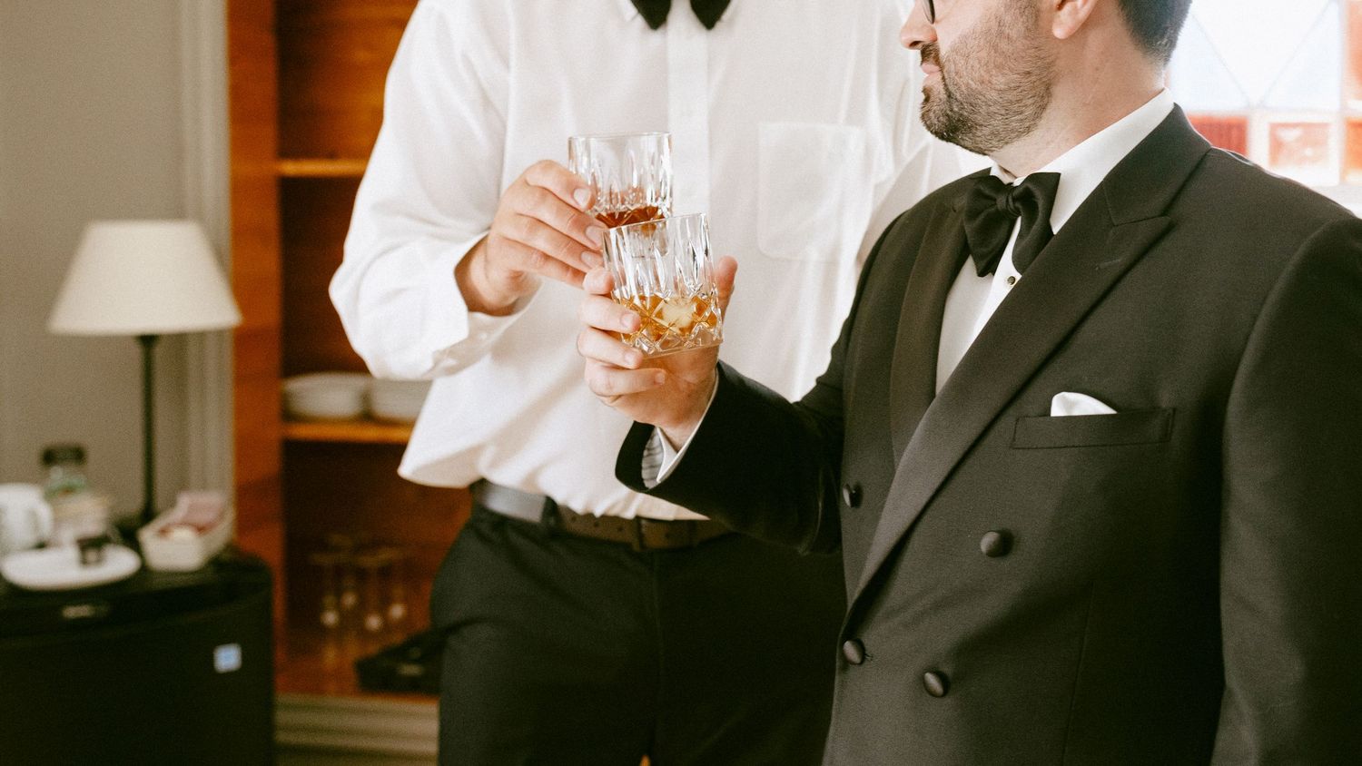 Two grooms in tuxedos toasting with whiskey glasses during wedding day preparations.