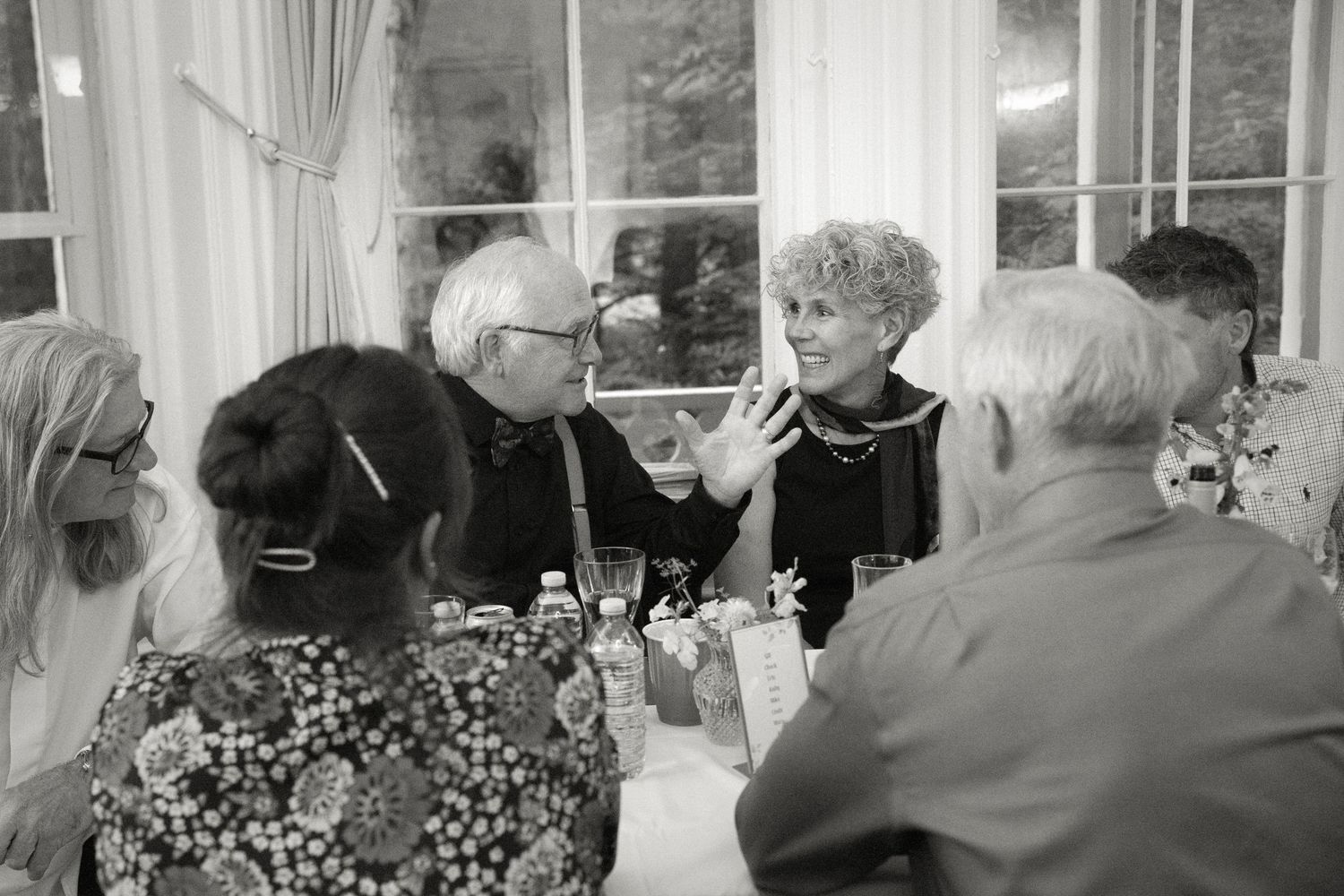 Elderly guests engaged in lively conversation at a wedding reception table in black and white.