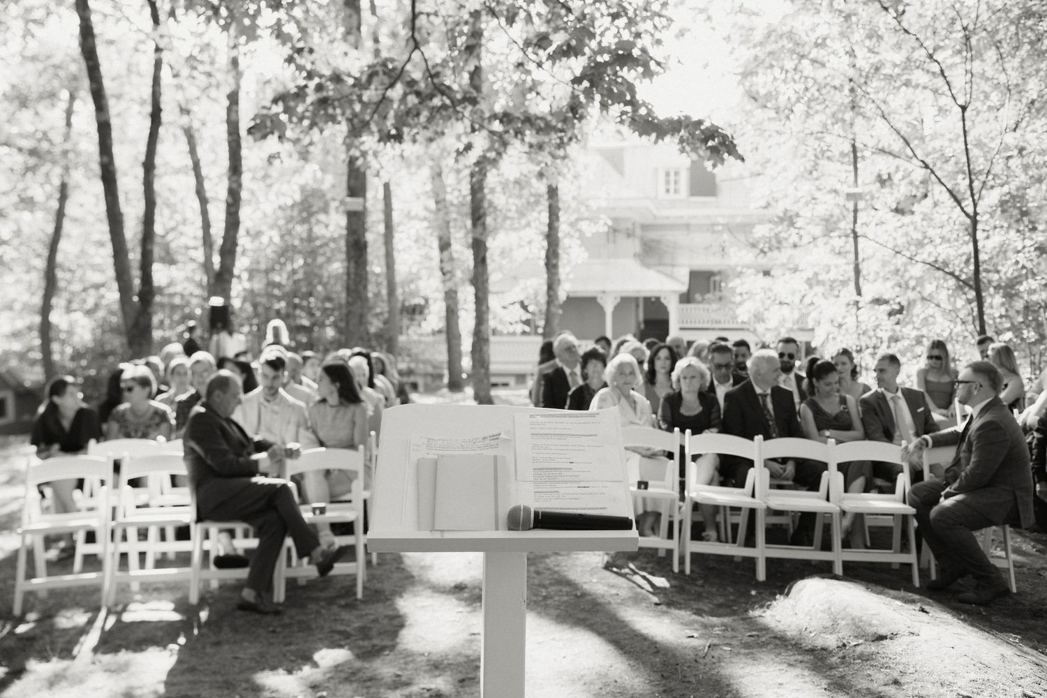 Guests seated in white chairs attend an outdoor wedding ceremony surrounded by lush trees.