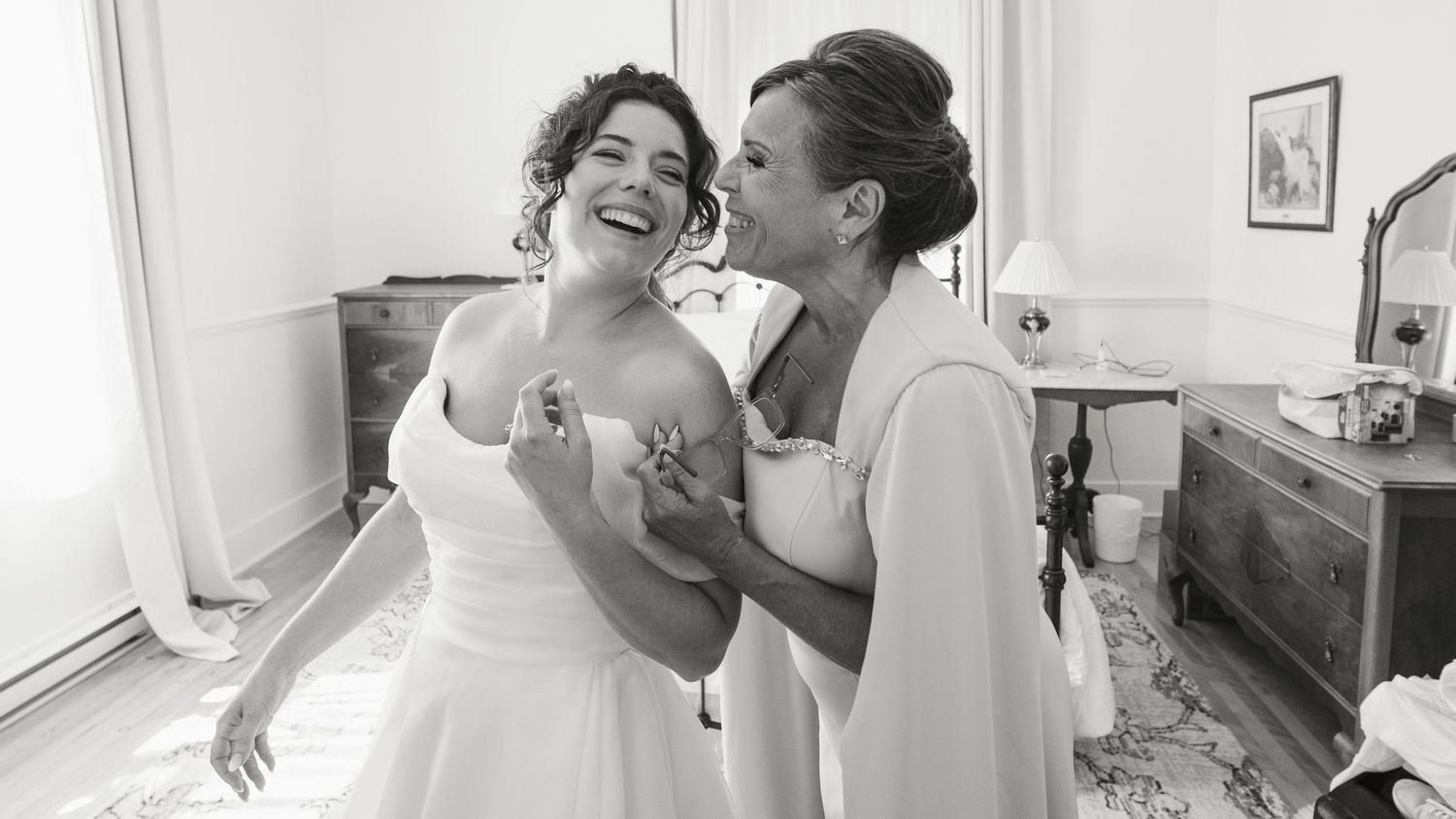 Two women laughing joyfully together, bride in white wedding dress being helped by companion, black and white.