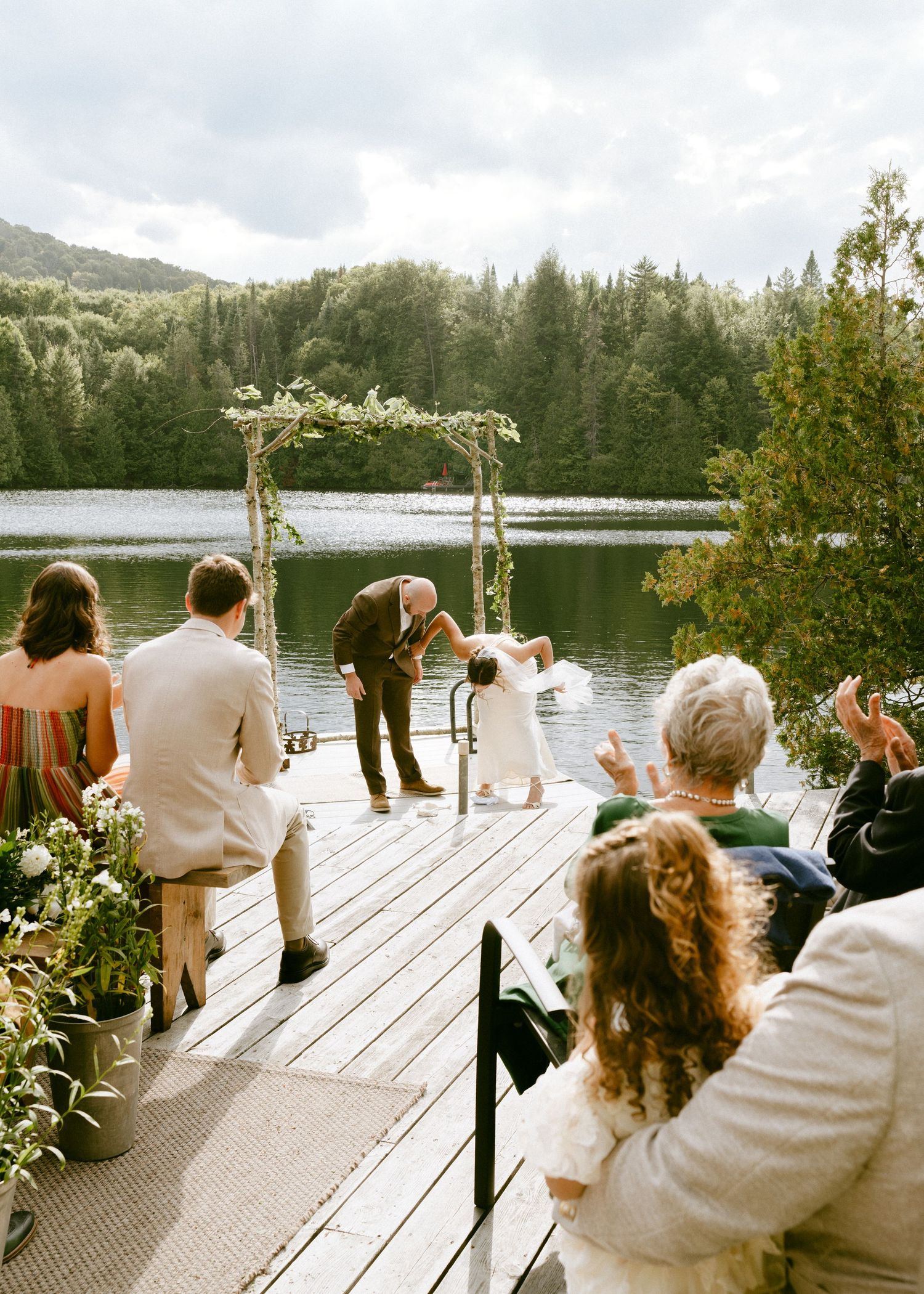 Wedding guests applaud as bride and groom share a dip kiss on a scenic lakeside dock.