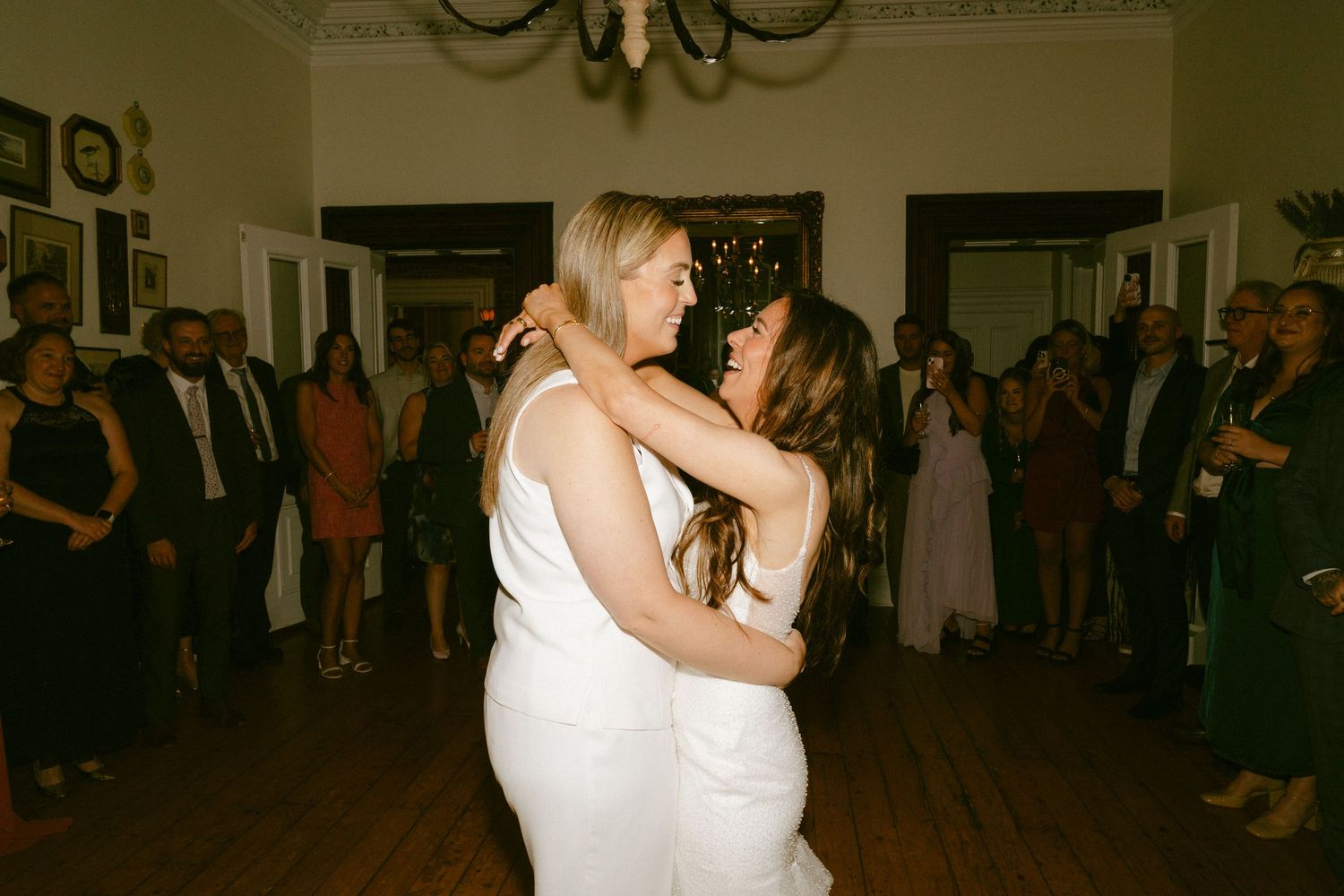 Two brides share their first dance embracing closely while wedding guests watch in a elegant venue.