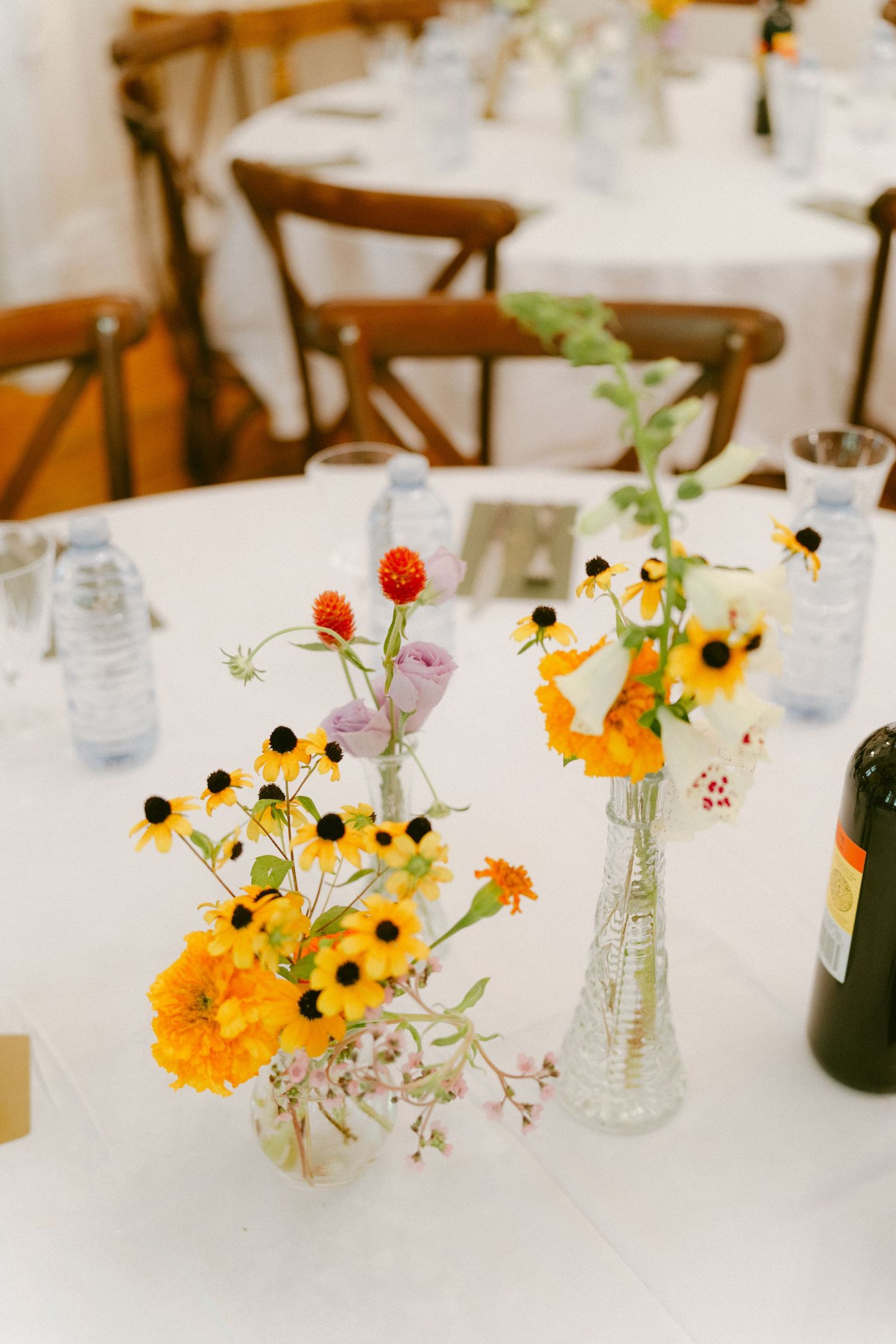 Bright yellow and orange wildflower centerpieces in glass bud vases on a white wedding reception table.