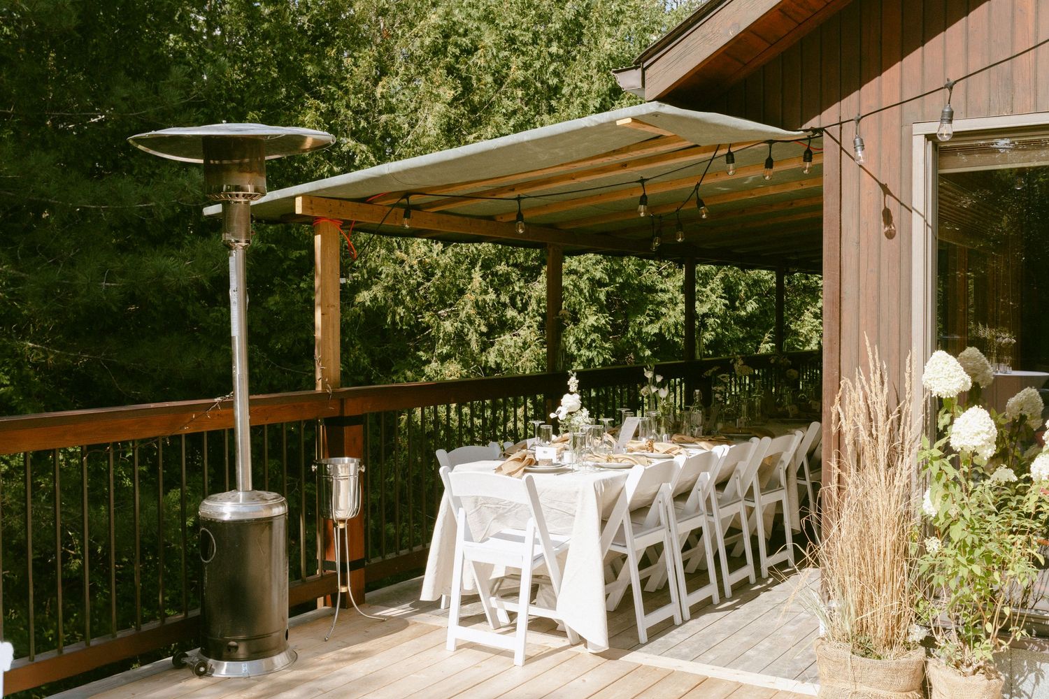 Outdoor wedding reception table with white chairs set on a wooden deck surrounded by lush green forest trees.