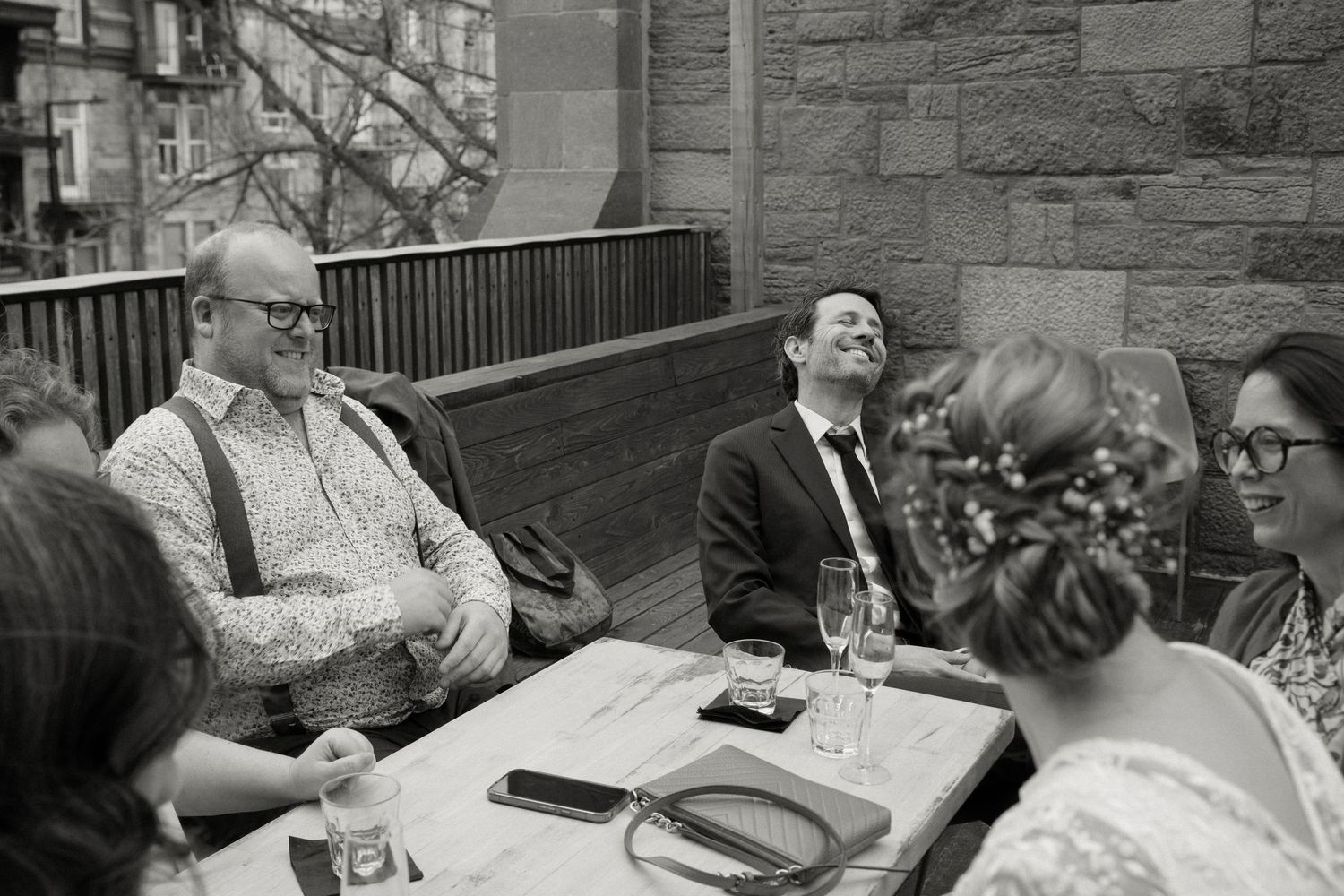 Black and white photo of wedding guests laughing and celebrating at an outdoor table during a reception.