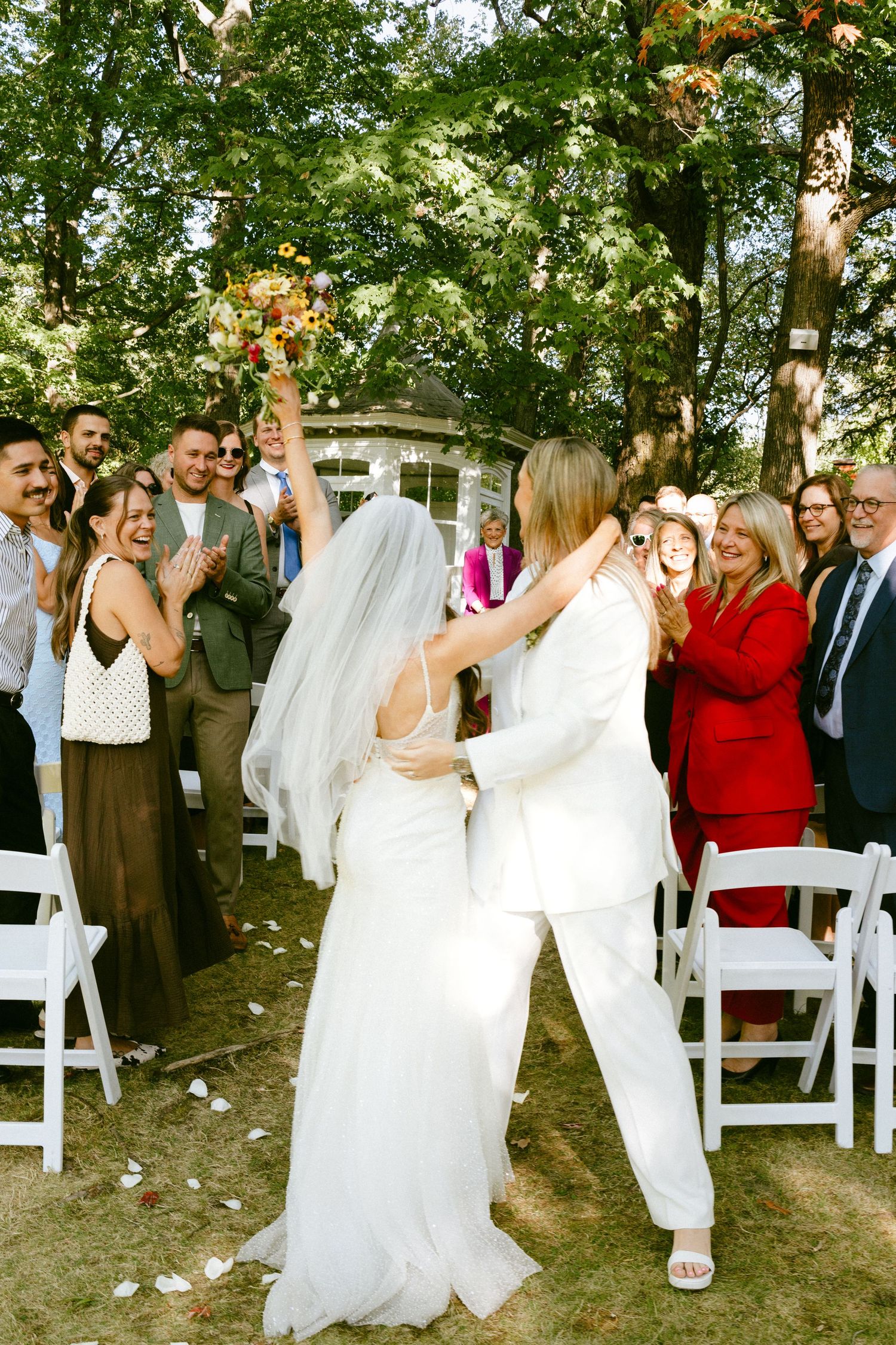 Two brides embrace and celebrate at the altar surrounded by cheering guests at an outdoor wedding.