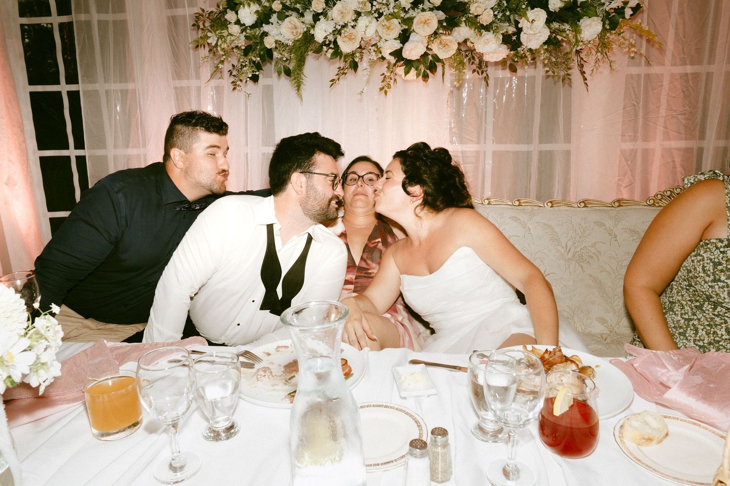 Bride and groom receiving kisses from wedding guests at reception table.