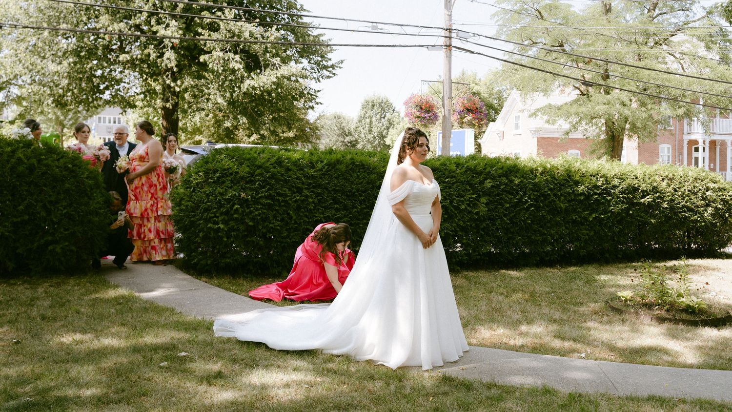 Bride in white off-shoulder gown with long train stands outdoors while bridesmaid adjusts her dress.