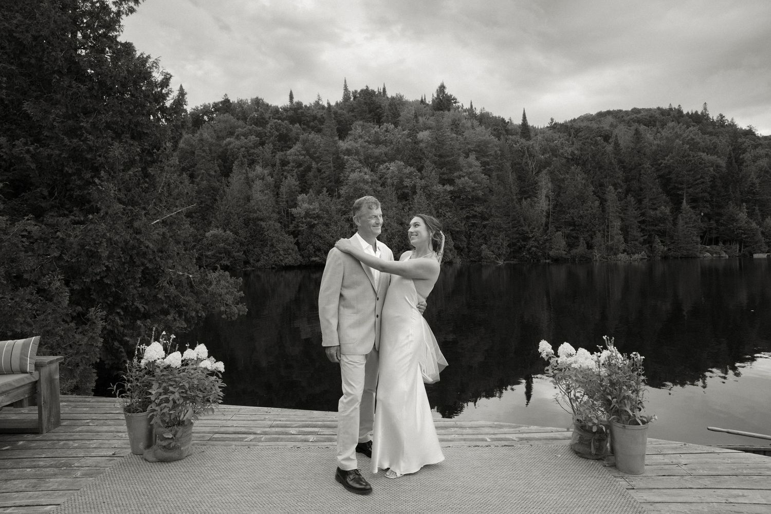 Bride and groom dancing on a lakeside dock with forested hills reflected in calm water, black and white photo.