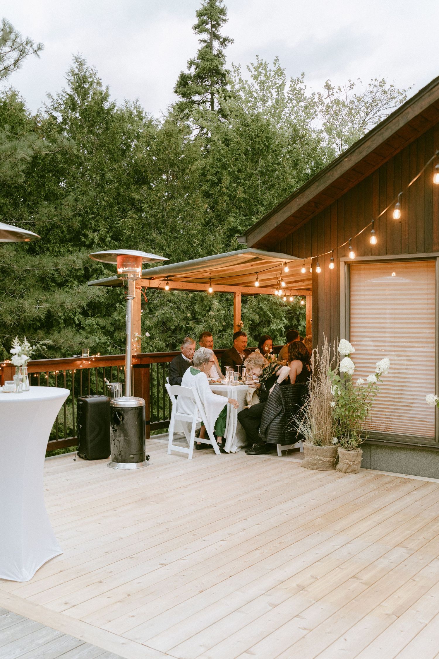 Small group of wedding guests dining at an outdoor deck table under string lights at dusk.