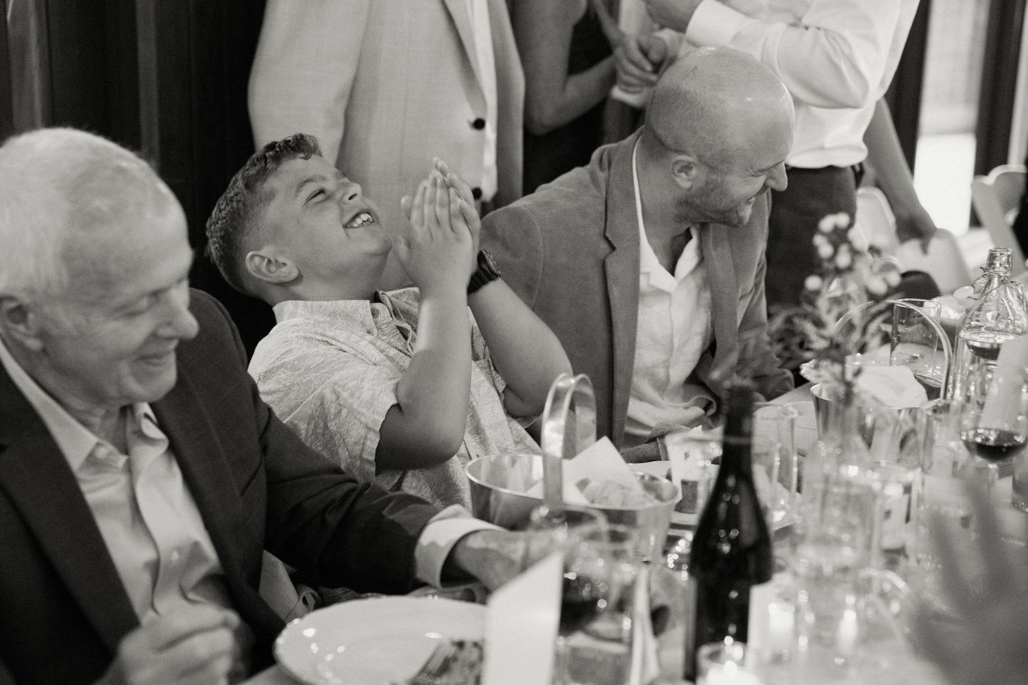 Black and white photo of a boy laughing joyfully at a wedding reception table surrounded by older guests.