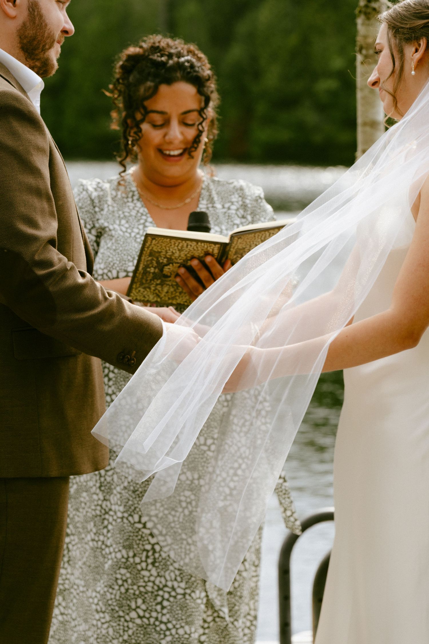 Smiling officiant reads from a book as bride and groom hold hands during their wedding vows.