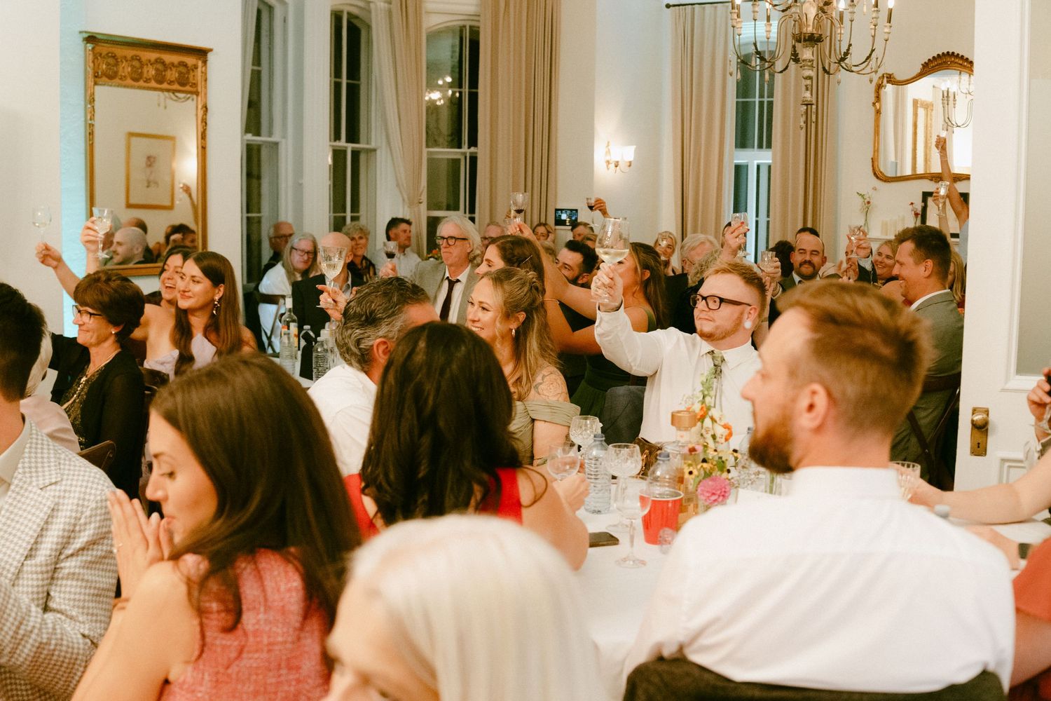 Wedding reception guests raise glasses in a toast inside an elegant white dining room with chandelier.