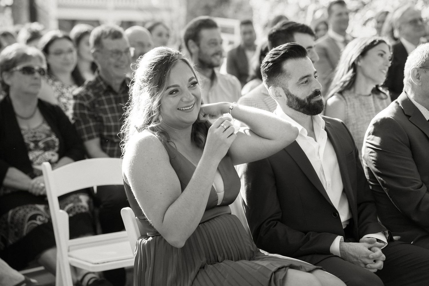 Black and white photo of smiling wedding guests seated during an outdoor ceremony.