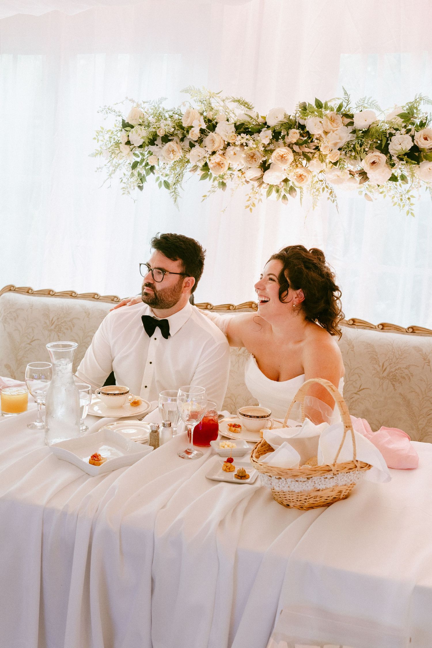 Bride and groom laughing together at sweetheart table beneath white floral centerpiece arrangement.