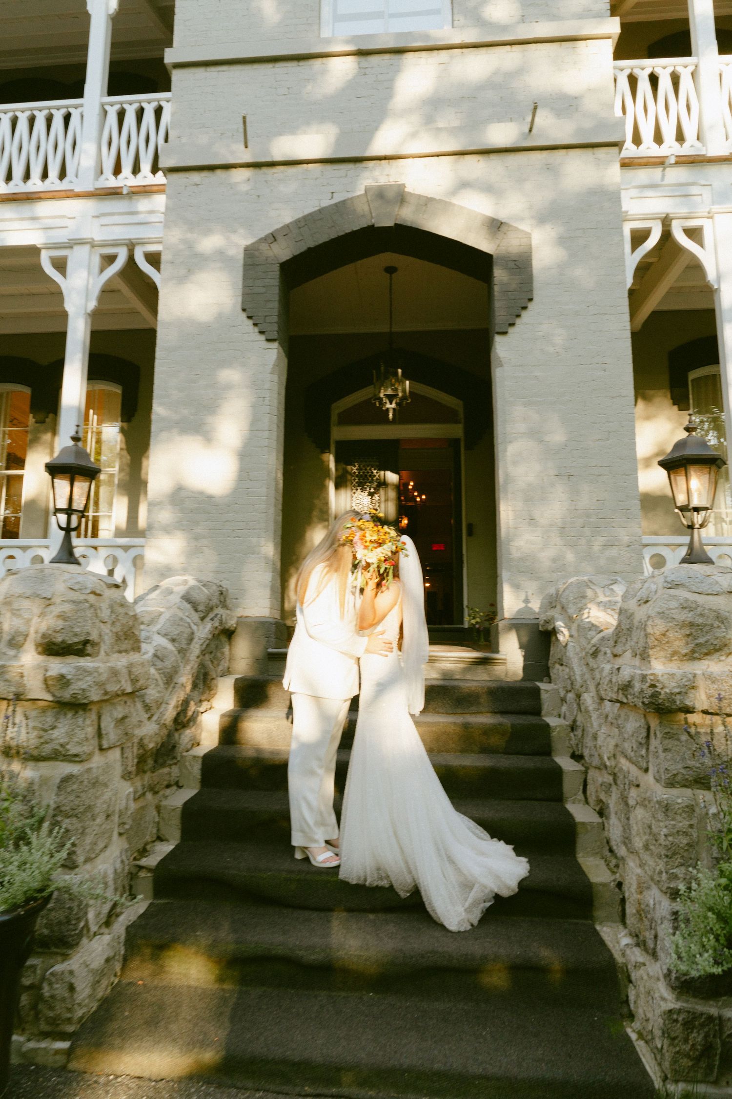Bride in flowing white gown walking up stone steps of an elegant historic mansion entrance.