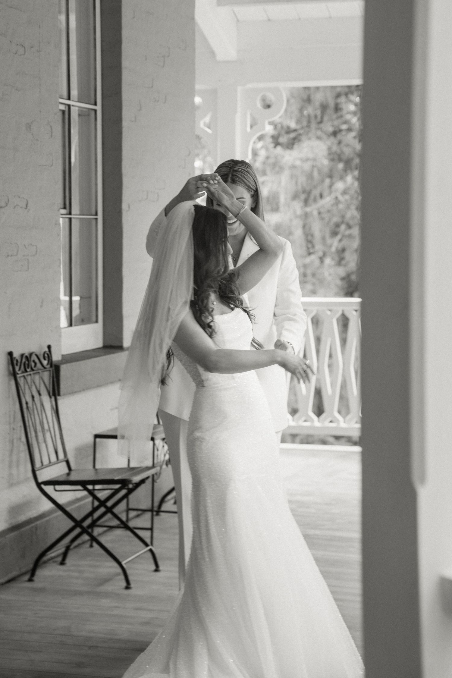 Two brides share tender embrace on porch in black and white wedding photo.