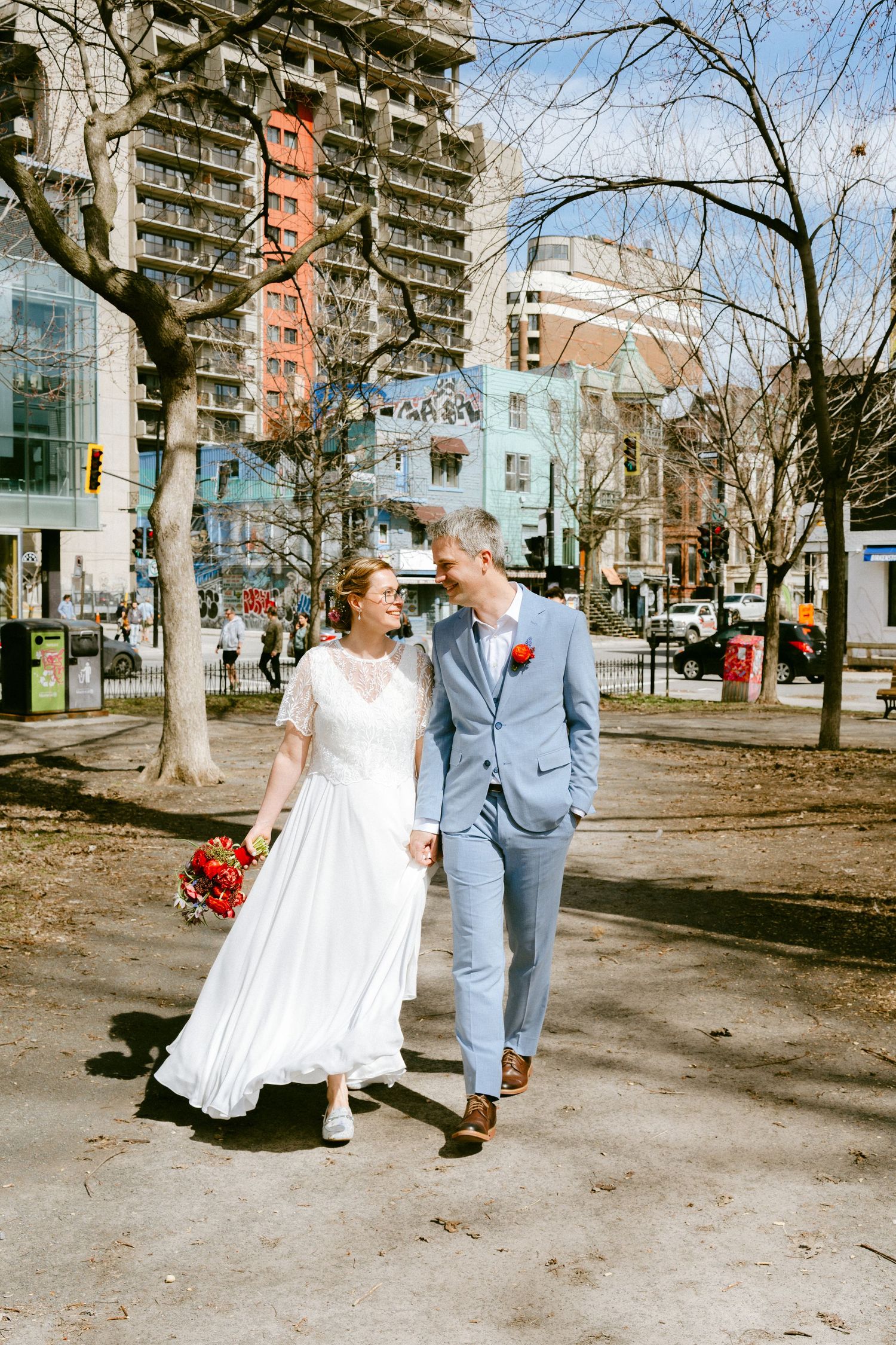 Smiling bride and groom walk through an urban park holding a red bridal bouquet.