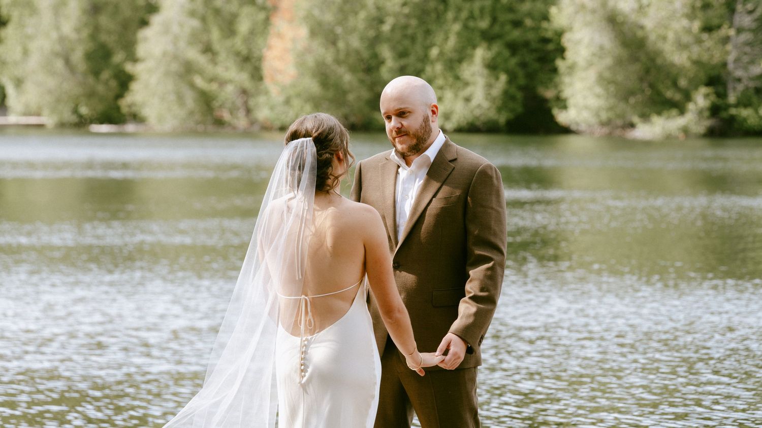 Bride in backless gown with veil holding hands with groom in brown suit beside a serene forest lake.