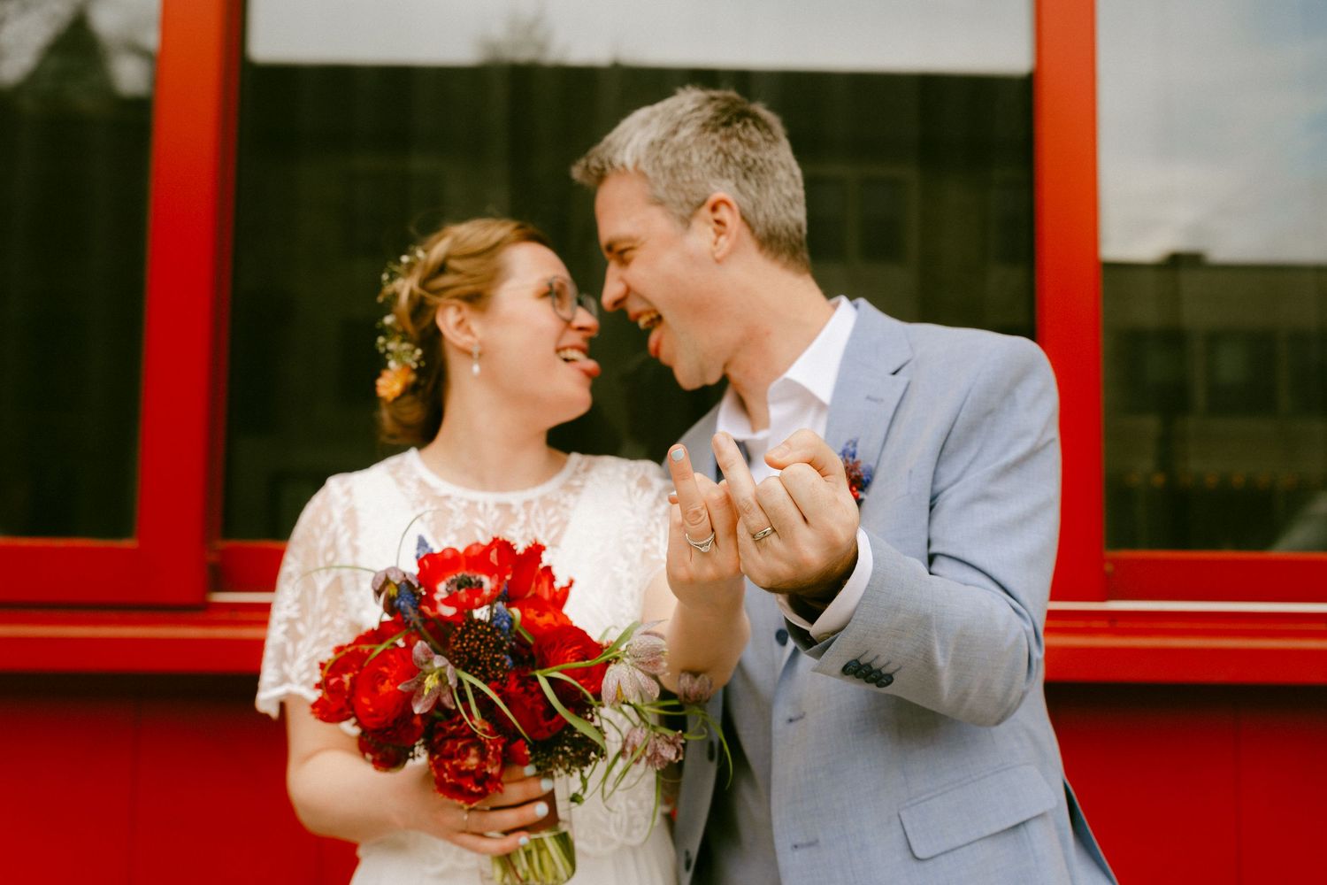 Bride and groom laughing together holding red poppy bouquet in front of red background.