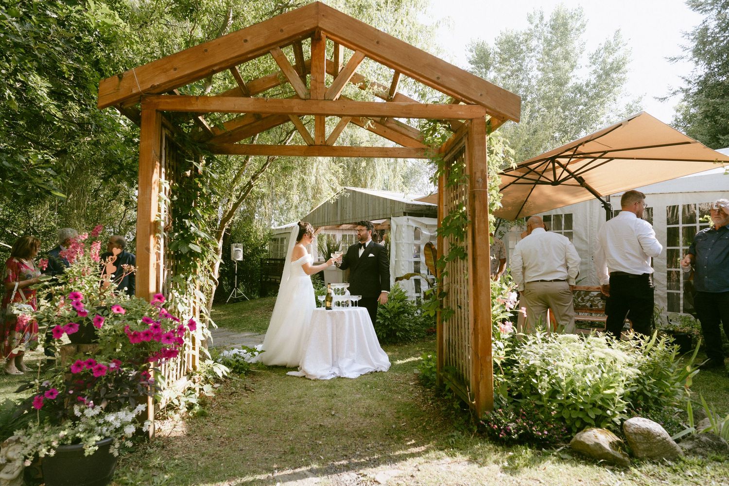 Bride and groom exchange vows under a wooden pergola surrounded by colorful flowers at outdoor wedding.