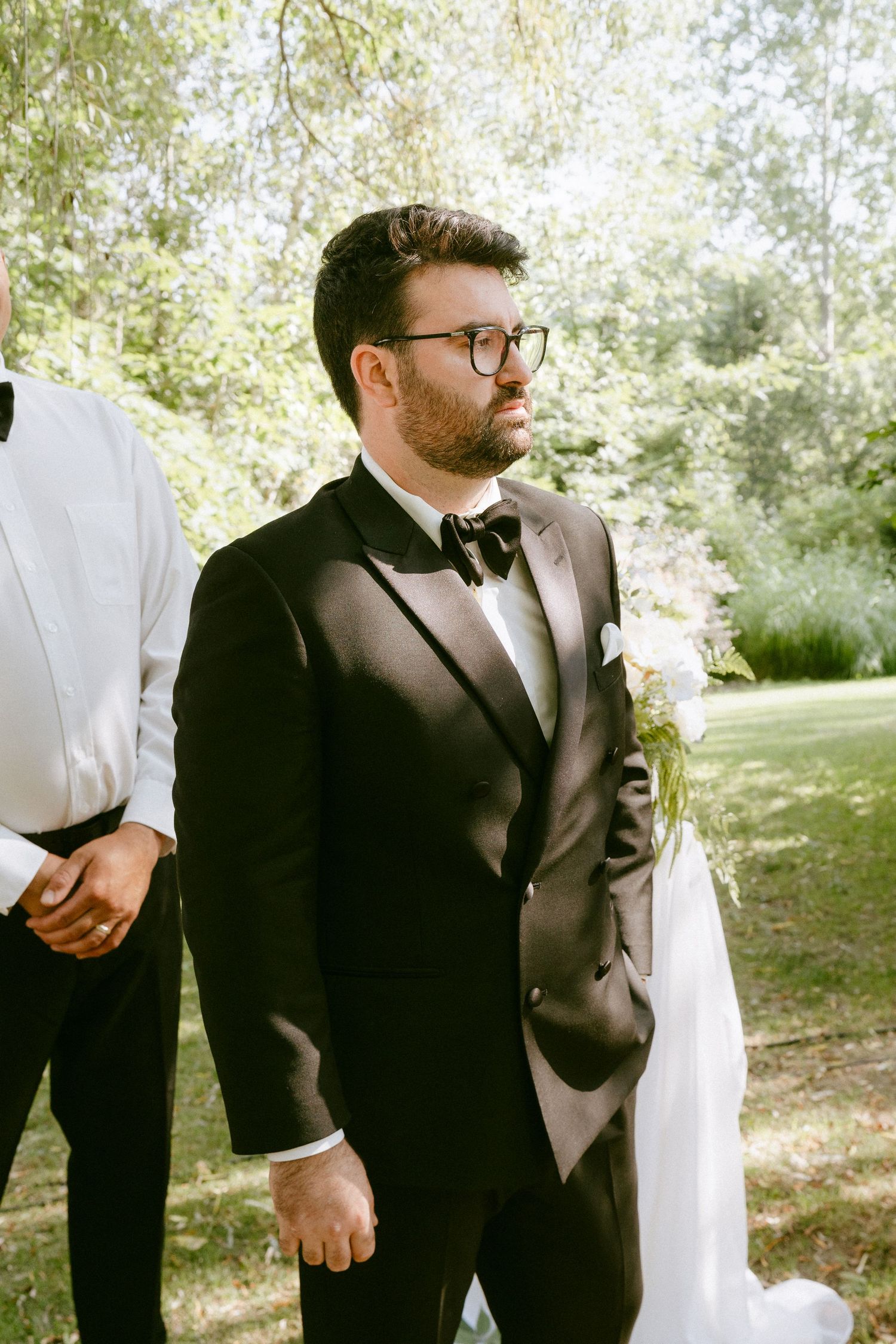 Groom in black tuxedo and glasses awaits bride at outdoor wedding ceremony.