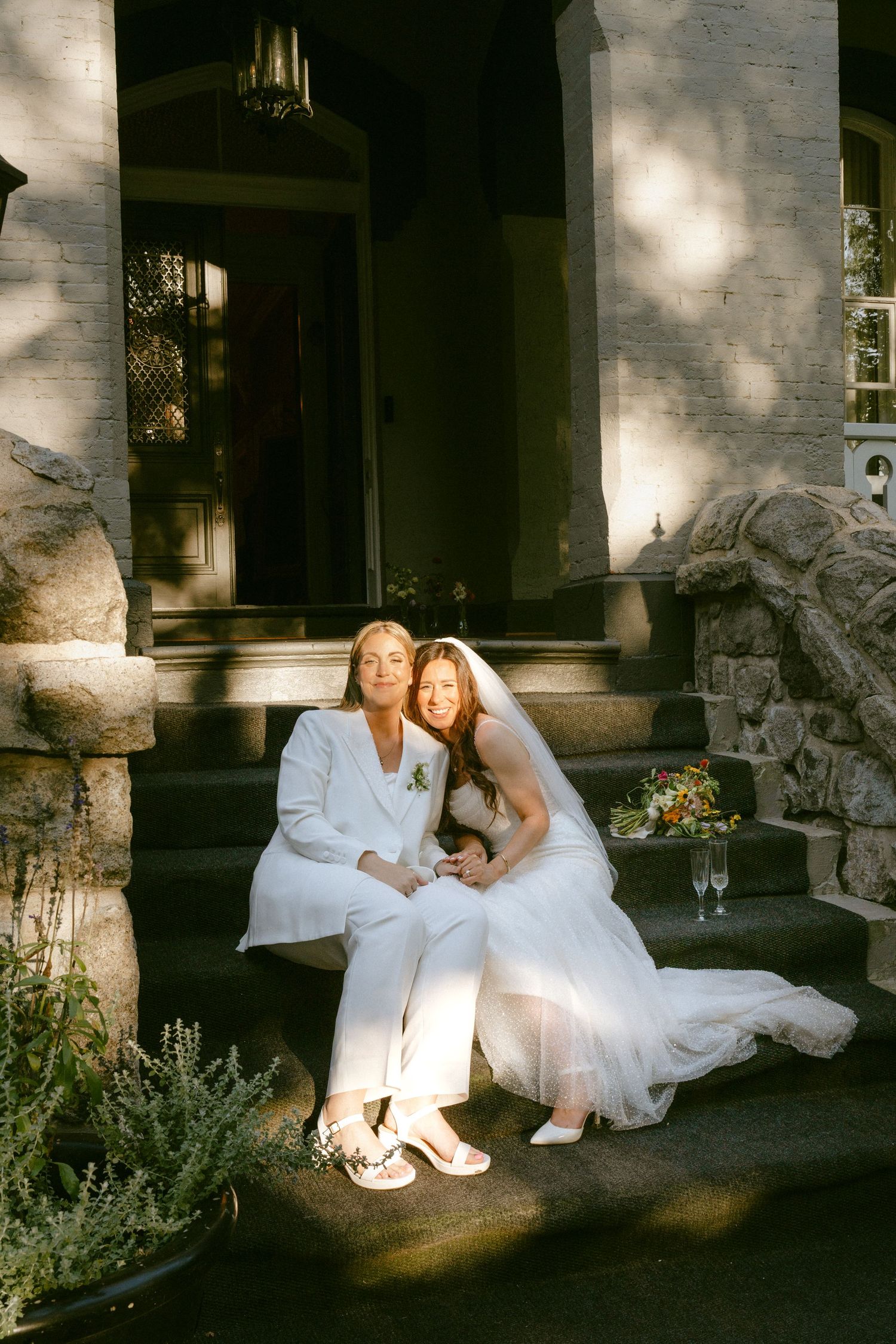 Two brides in white wedding attire sitting together on stone steps of a historic estate.