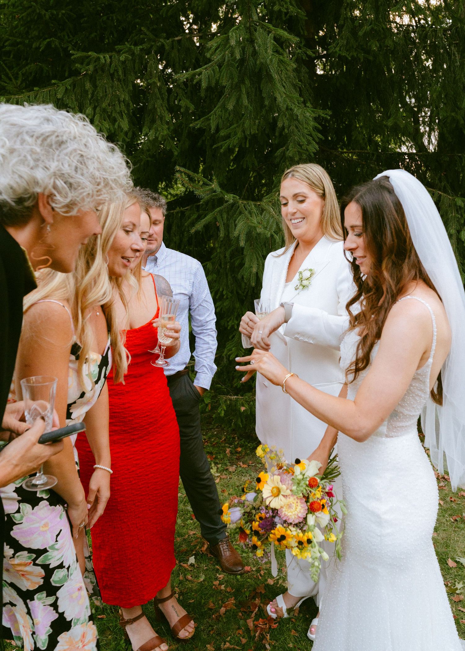 Bride in veil holding colorful bouquet shows wedding ring to smiling guests at outdoor reception.