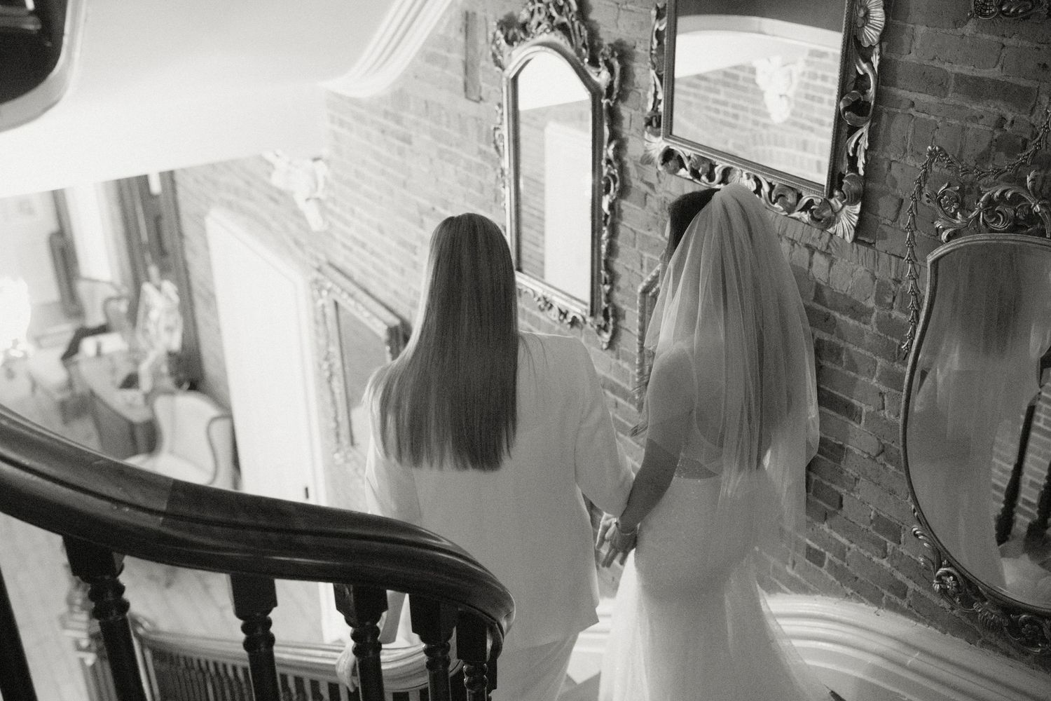 Two brides hold hands on ornate staircase in black and white wedding photo.